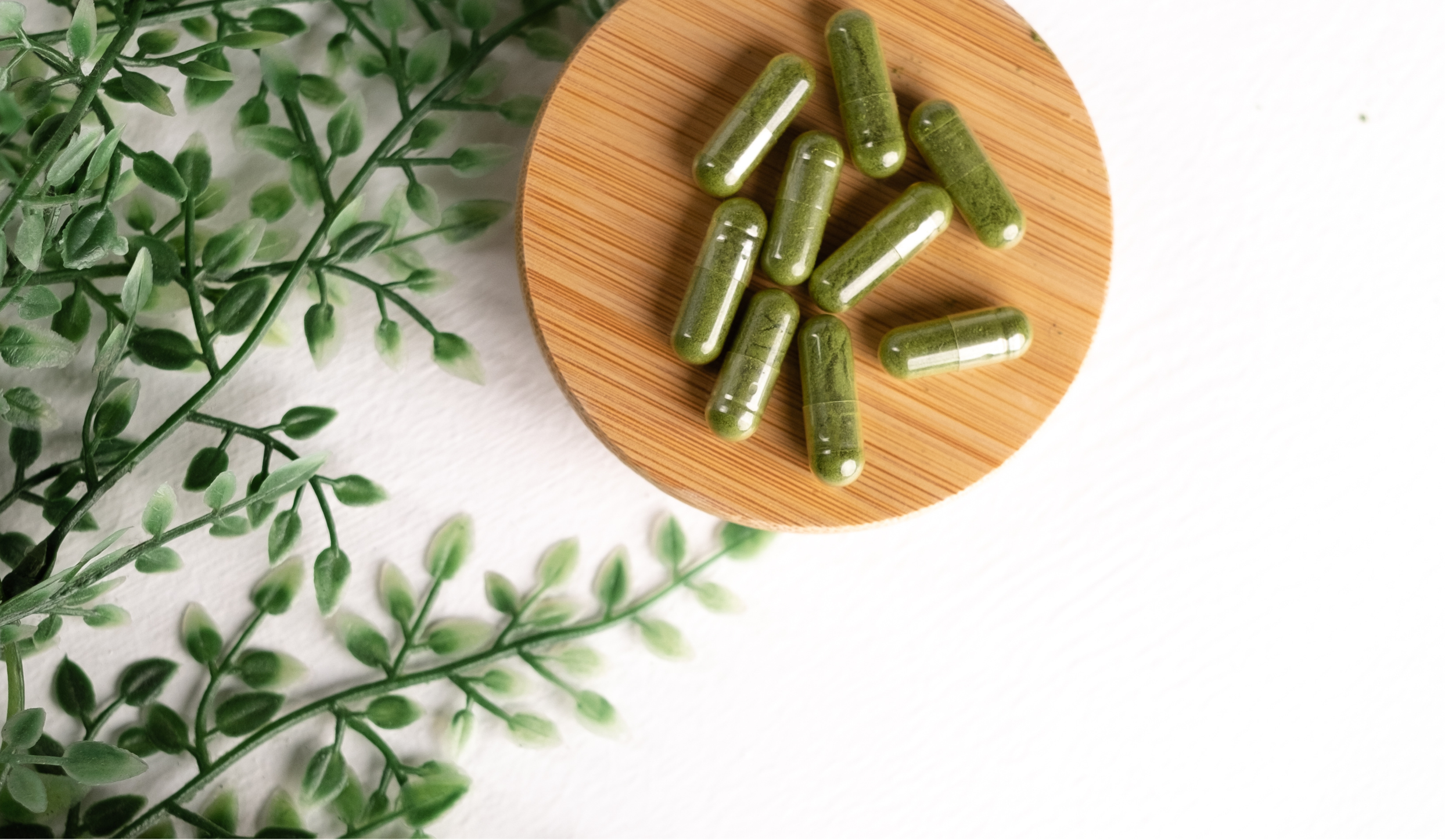 Green herbal supplement capsules on a round wooden dish, with green faux plant leaves beside it on a white surface.