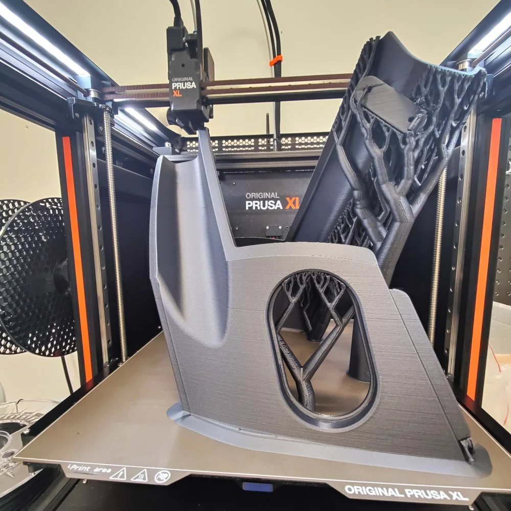3D-tulostin tulostamassa mustaa muoviosaa, jossa on pyöreä aukko ja alakulmassa teksti 'ORIGINAL PRUSA XL'.