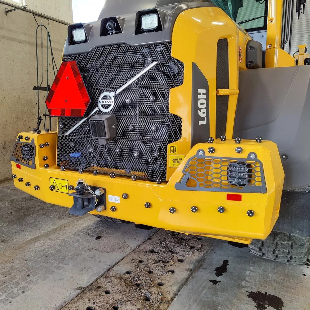 Koneen etupuoli, jossa näkyy Volvo-logolla varustettu moottori ja varoitusmerkki, ja punainen kolmiomerkki. Keltaiset ja mustat osat, useita pienikokoisia metallipalloja ja sähkötarvikkeita näkyvissä.