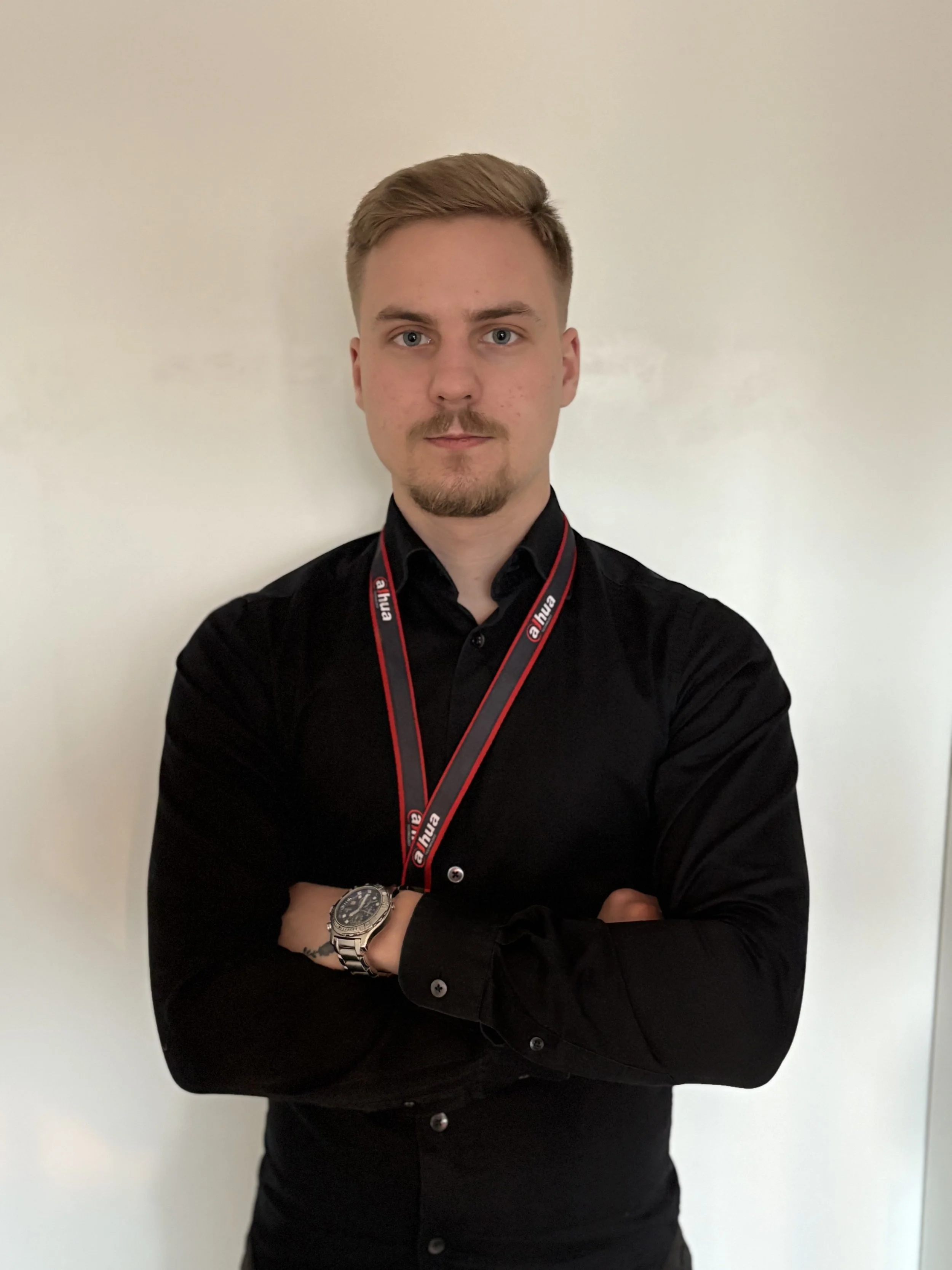 Person wearing a black shirt with a lanyard, posing with arms crossed against a plain background.