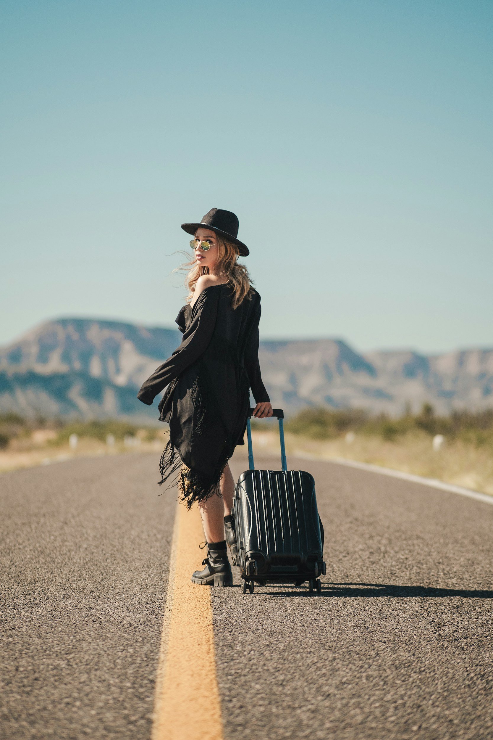 Woman standing on an empty road in a desert, pulling a black suitcase, wearing a black hat, black dress, and sunglasses, with mountains in the background.