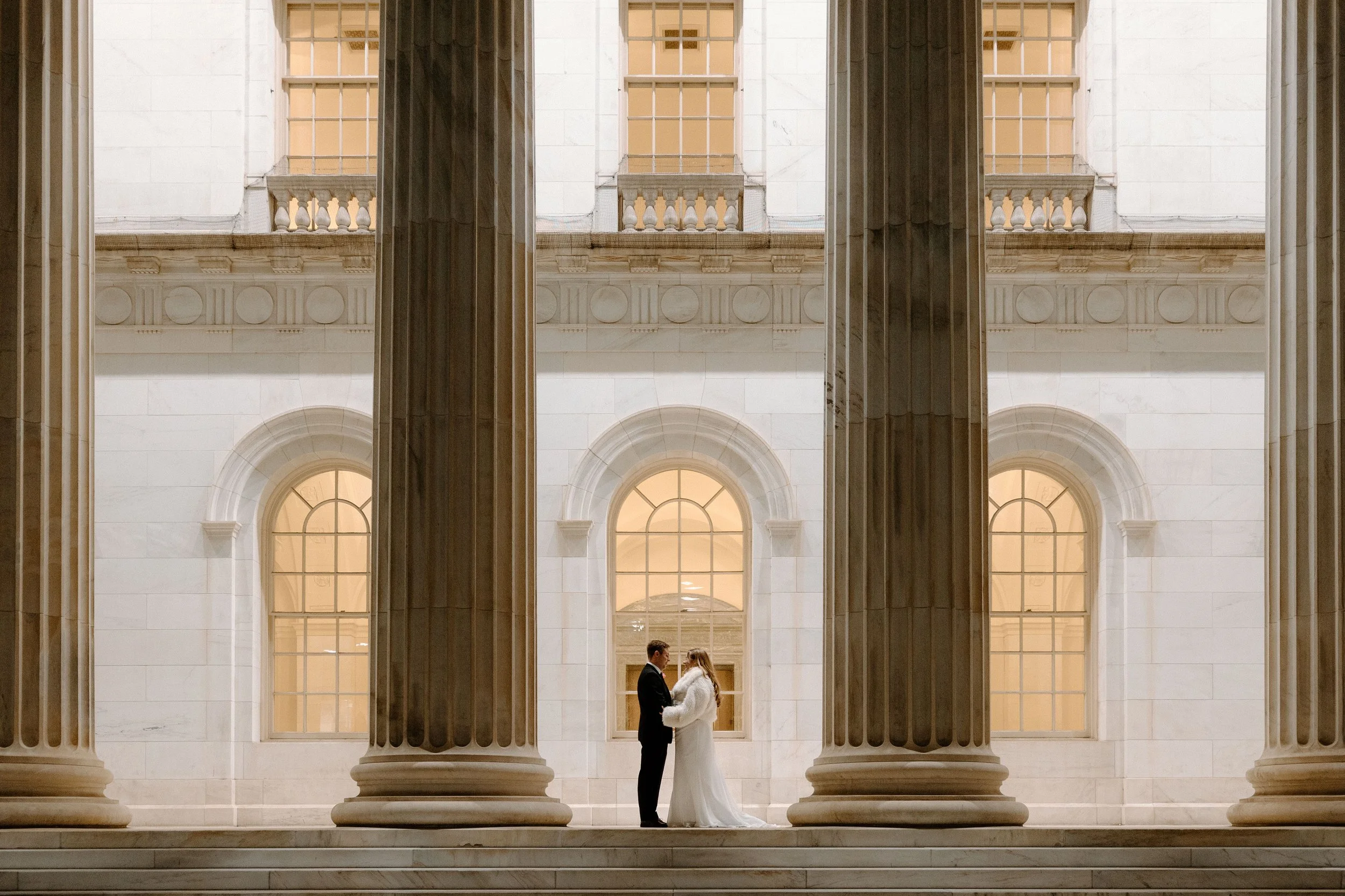 A bride and groom standing face-to-face in front of large white marble columns inside a grand neoclassical building, with tall arched windows behind them, in a formal setting.