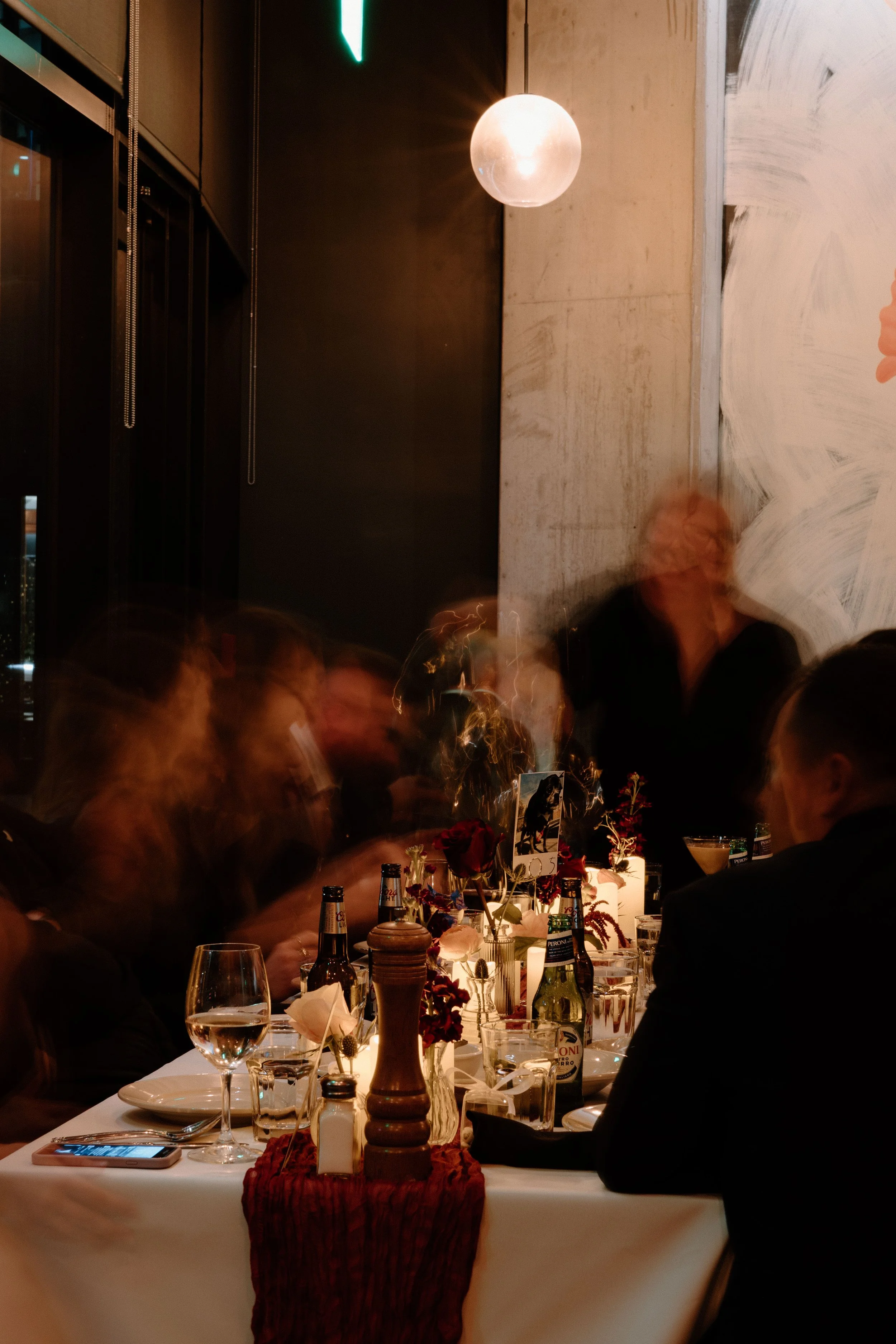 People sitting around a decorated dinner table with flowers, candles, and various beverages, in a dimly lit restaurant with modern decor and a round hanging light fixture.