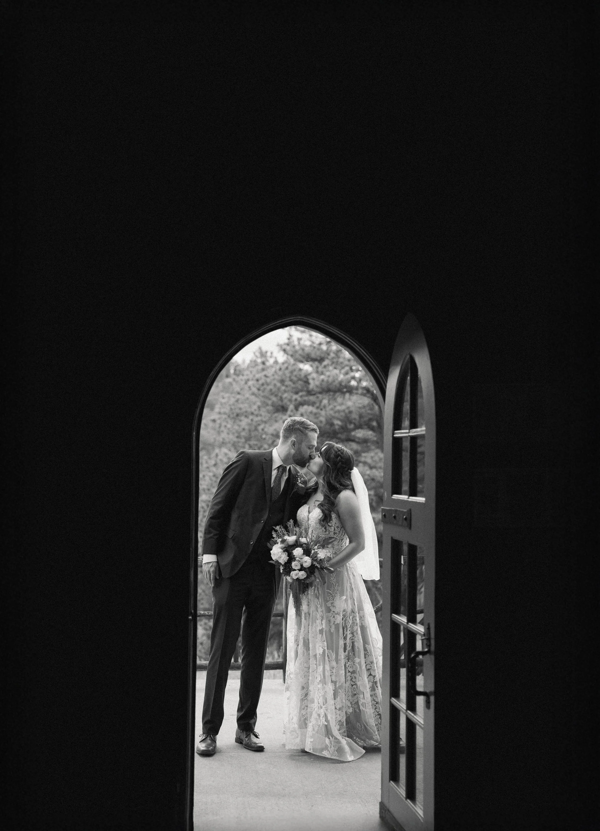 Black and white photo of a bride and groom kissing in a doorway, with trees visible outside.
