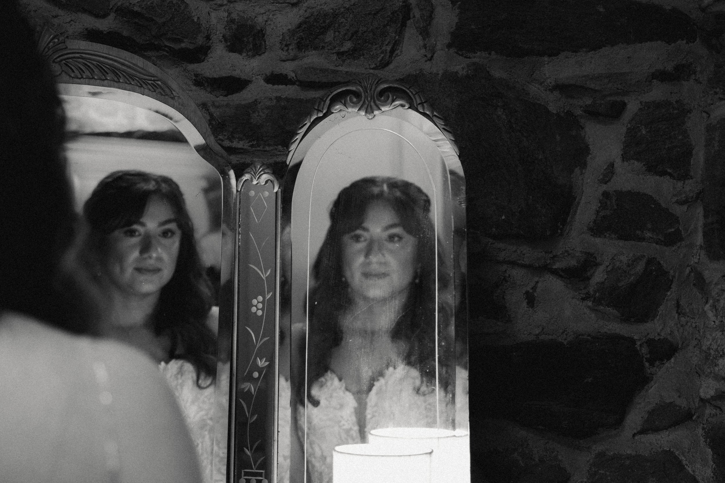 A woman with dark, wavy hair looking into a mirror with decorative framing, surrounded by a textured stone wall.