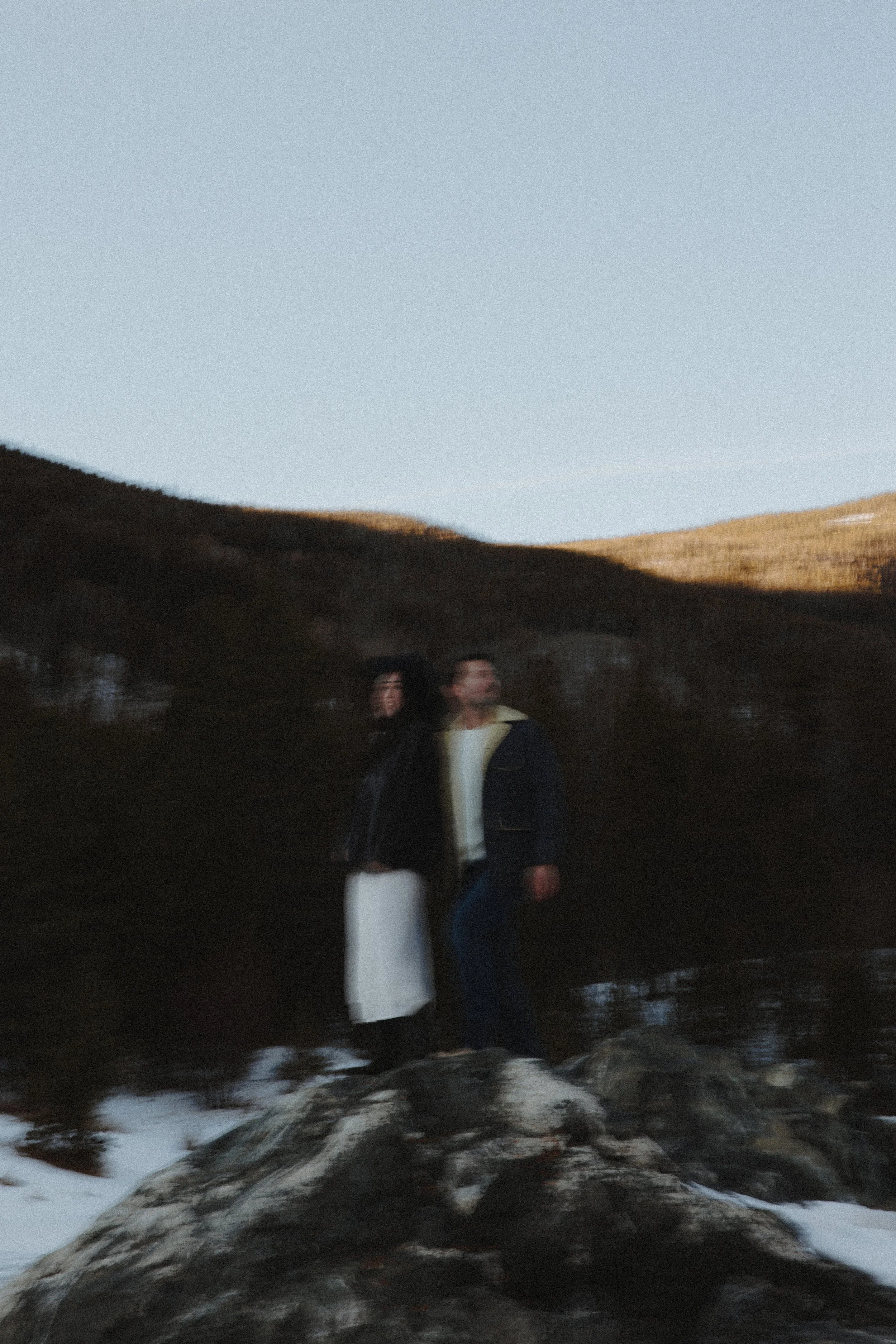 Two people standing on rocks near a body of water with mountains and a clear sky in the background, during what appears to be late afternoon or early evening.