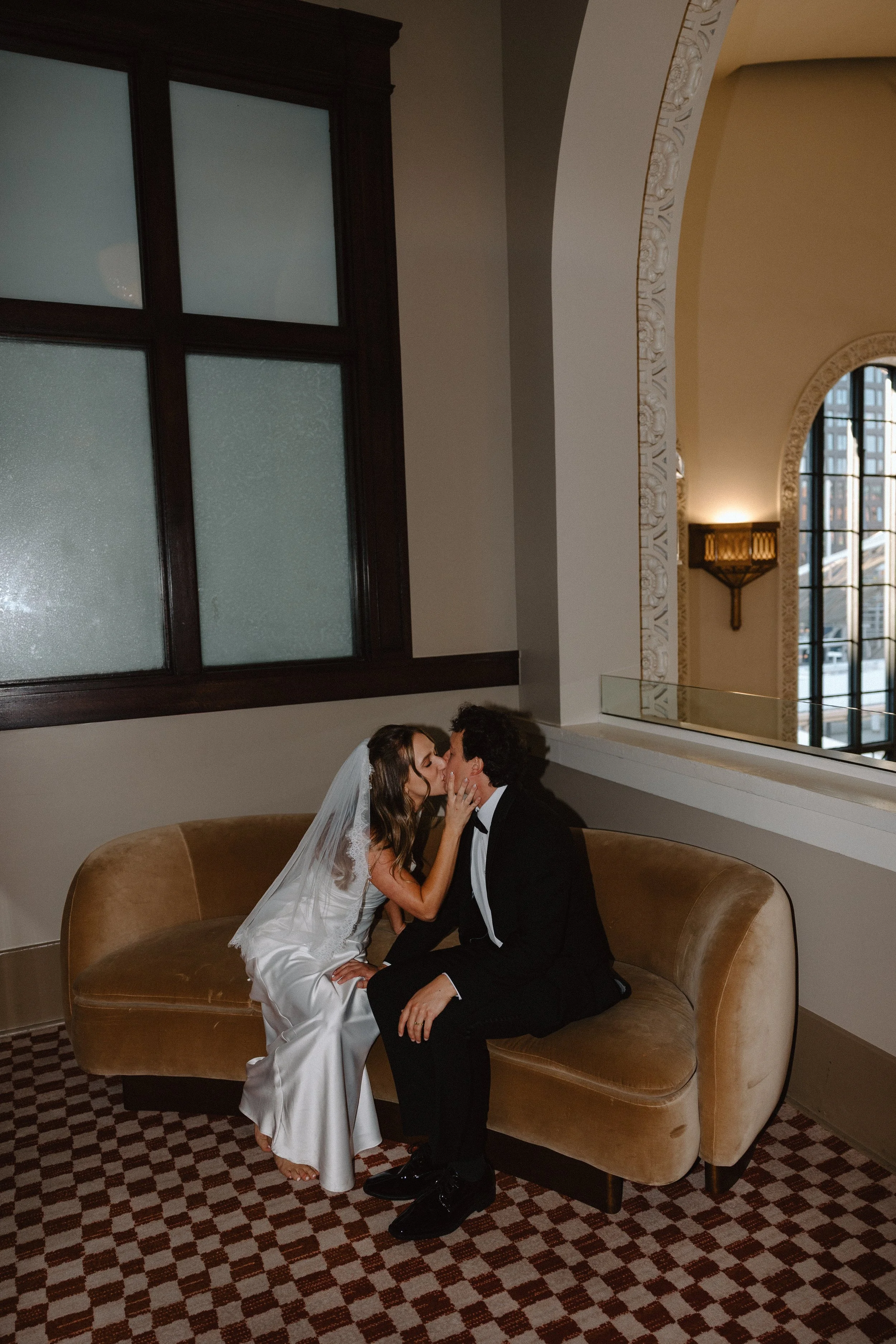 A bride and groom seated on a curved brown couch, kissing each other indoors near large windows with decorative frames. The bride is wearing a white wedding dress with a veil, and the groom is dressed in a black tuxedo with a bow tie. The room has ornate wall decorations and a checkered carpet.
