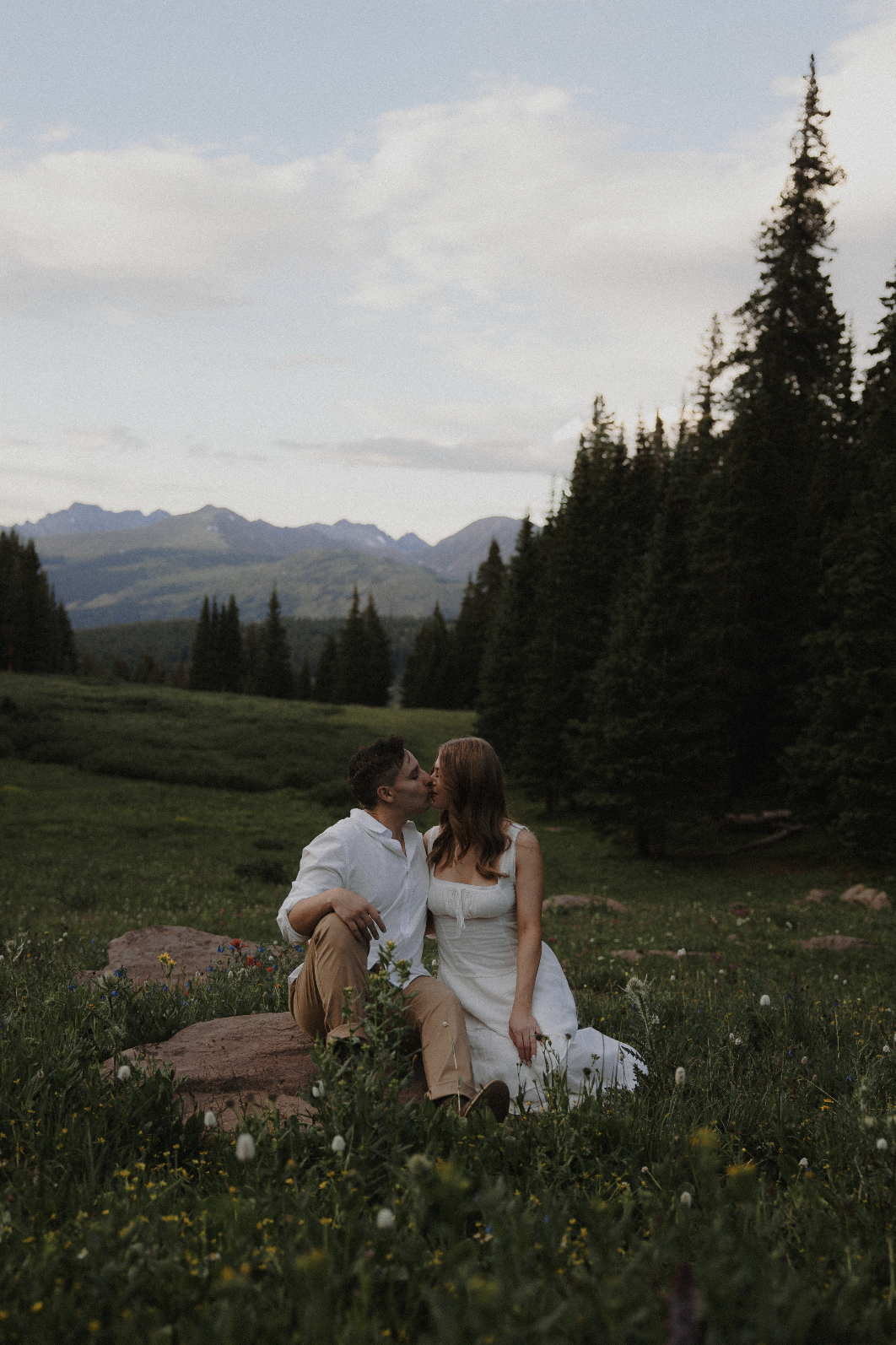 A couple kissing while sitting on a rock in a grassy field with wildflowers, surrounded by tall pine trees and mountains in the background during late afternoon or early evening.