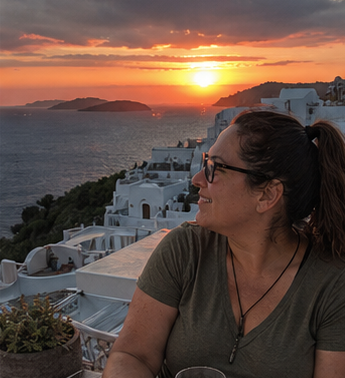 A woman wearing glasses and a green shirt sitting outdoors during sunset, overlooking a white coastal town and ocean.