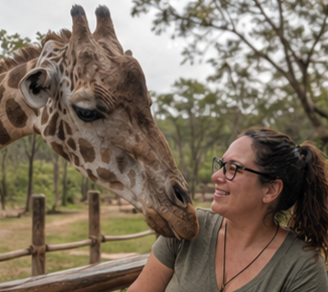 A woman with glasses smiling at a giraffe in an outdoor zoo setting.