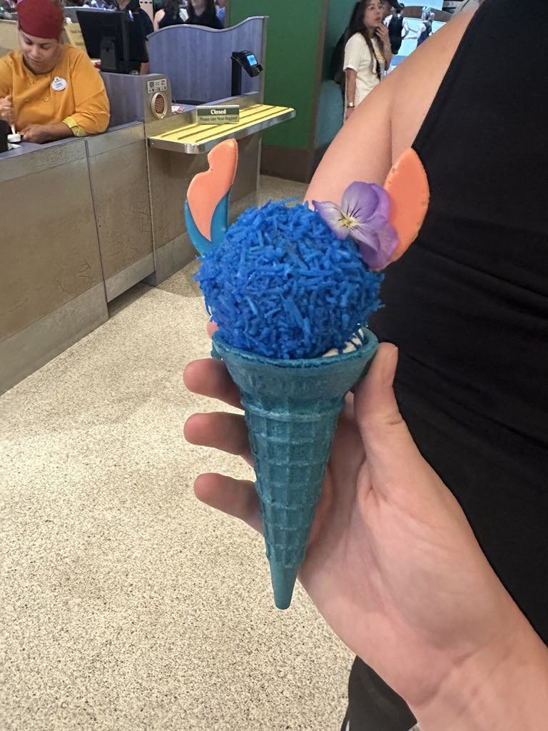 A person holding a cone with a scoop of bright blue shredded ice or candy, decorated with unicorn horn and ears made of fondant or paper, and a purple flower.