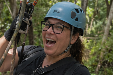 Woman wearing a blue helmet and glasses smiling during zip-lining in a forest.