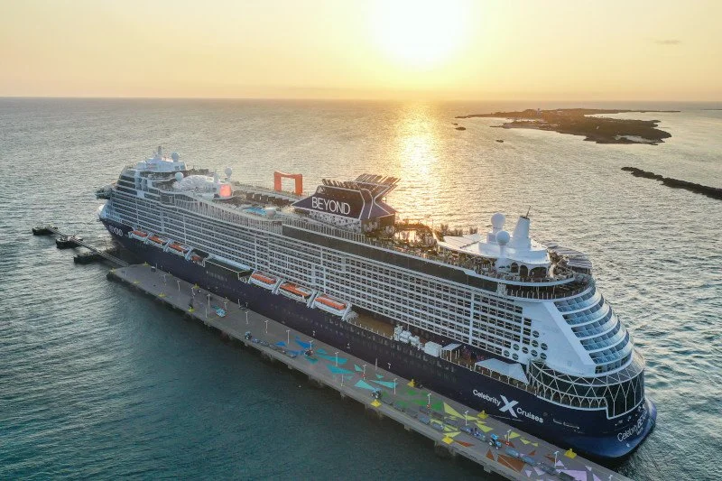 A large cruise ship docked at a pier during sunset with the ocean and small islands in the background.