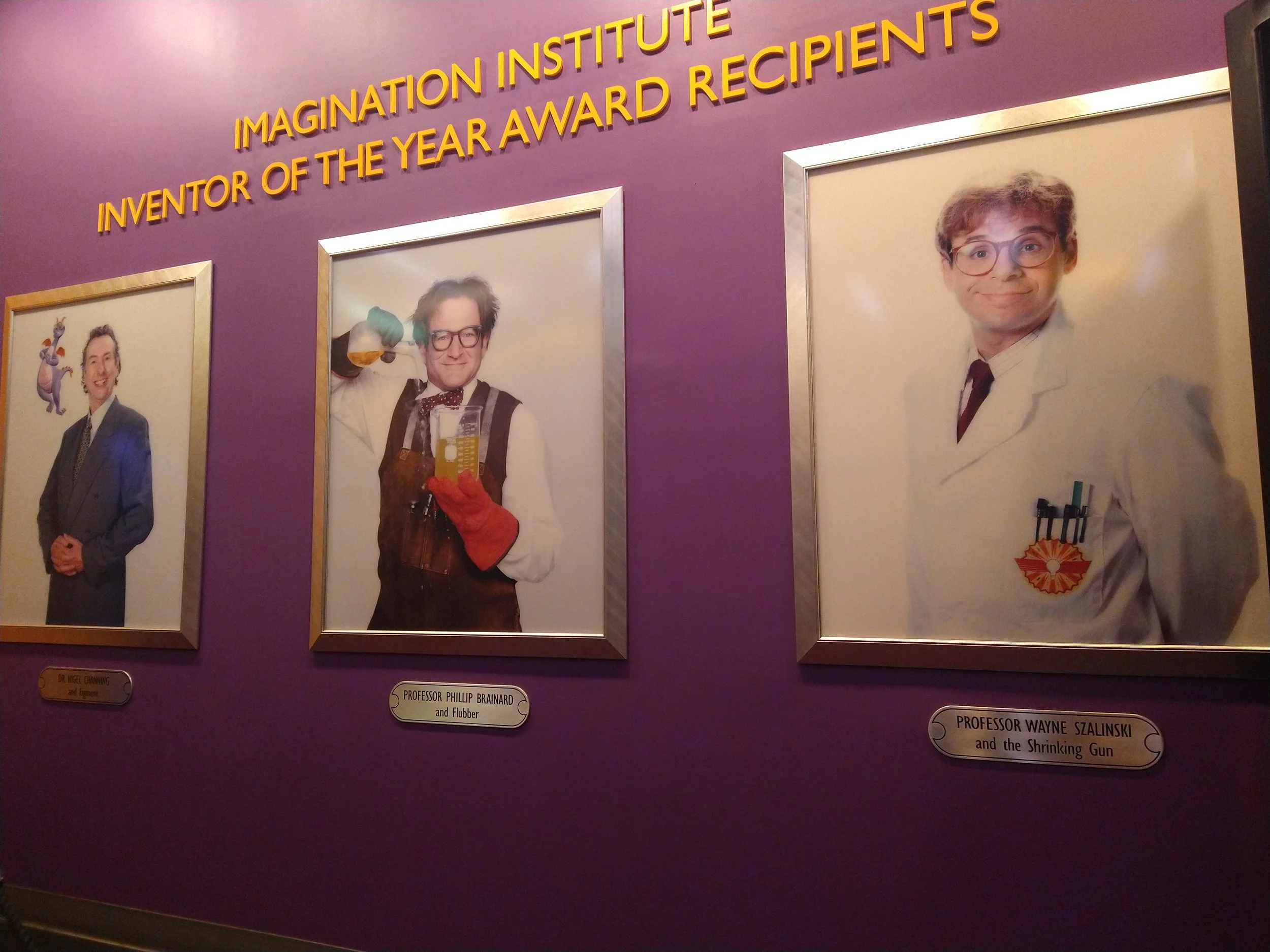 Photographs of three men on a purple wall with yellow text above them that reads "Imagination Institute, Inventor of the Year Award Recipients." The first man on the left is smiling and wearing a suit. The middle man is dressed as a scientist, holdin
