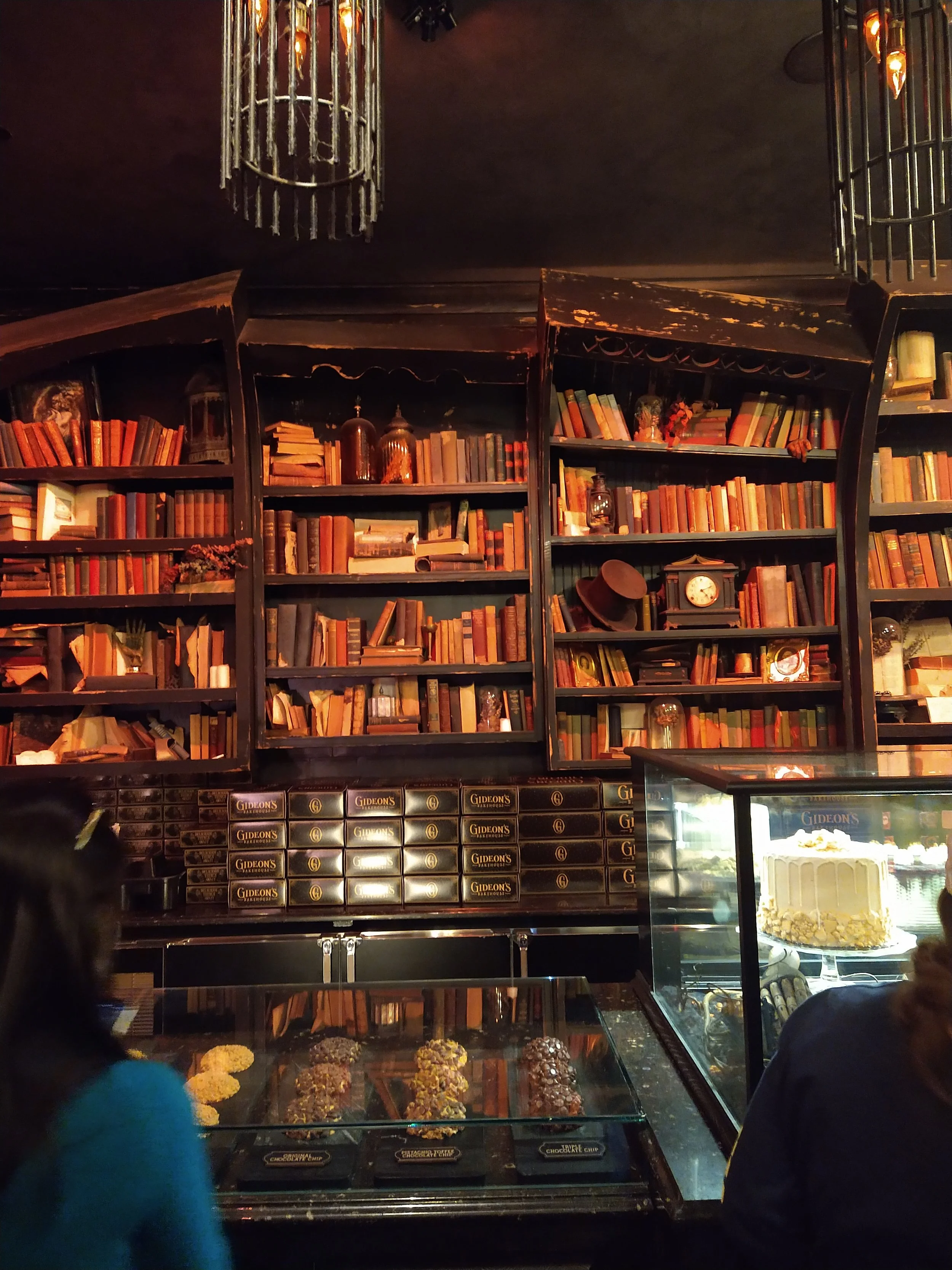 Interior of a bakery or cafe with shelves filled with books and vintage decor, and a display case with assorted chocolates or desserts, along with a cake in a glass case.