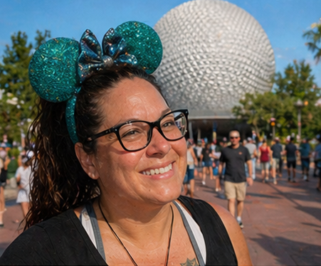 Woman wearing glasses and glittery teal Mickey Mouse ears in a theme park with the Spaceship Earth attraction in the background.