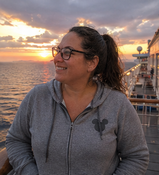 A woman wearing glasses and a gray hoodie with a Mickey Mouse logo, smiling and looking off to the side, on a ship at sunset.