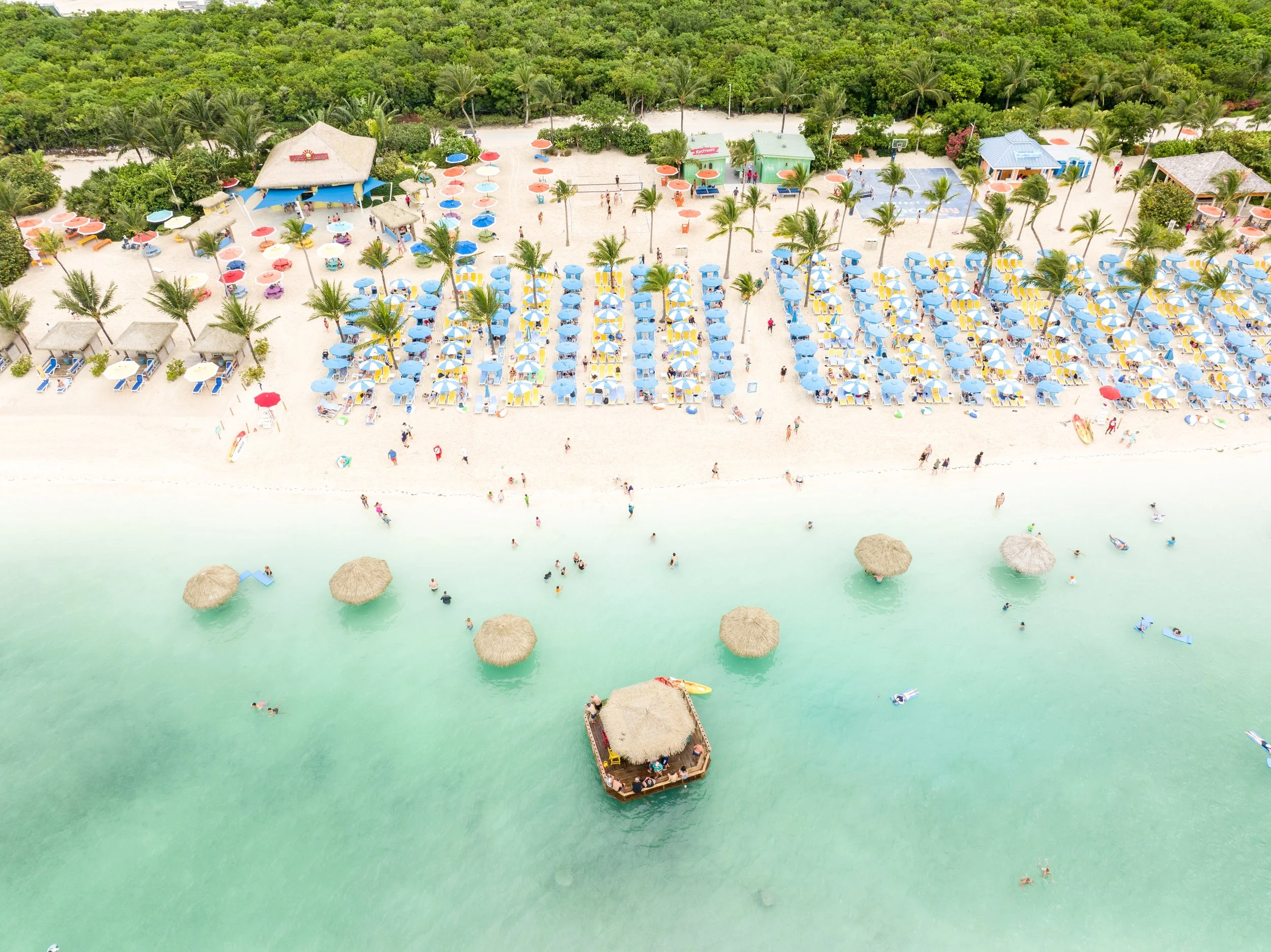 Aerial view of a tropical beach with colorful umbrellas, lounge chairs, cabanas, palm trees, and a tiki bar in turquoise water with people swimming and relaxing.