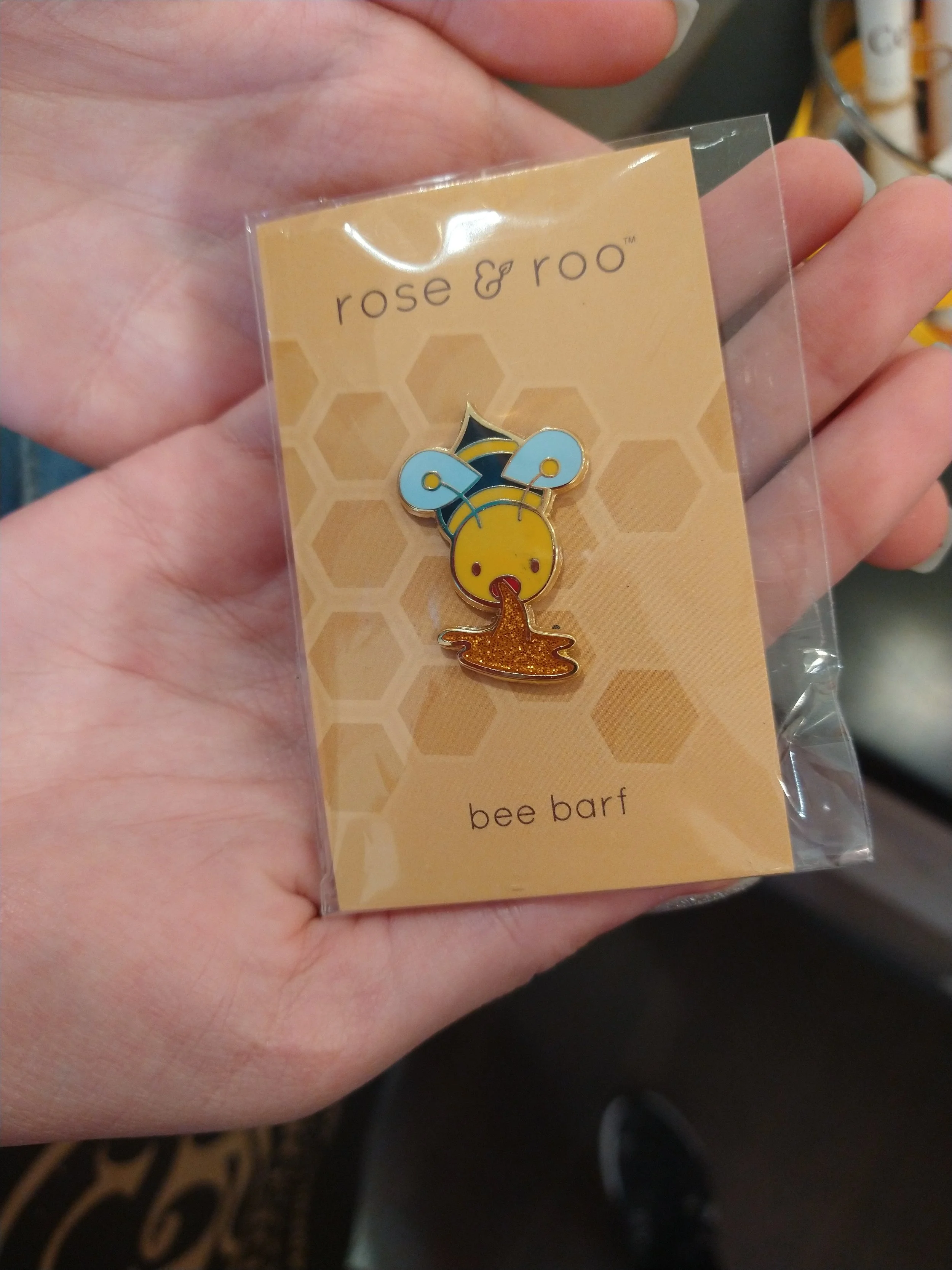 A hand holding a pin badge with a bee design, featuring a yellow face, blue wings, and a brown glittery stinger. The badge is attached to a cardboard backing with a honeycomb pattern and the text "rose & roo" and "bee barf."