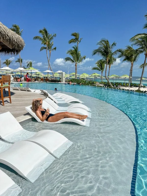 A woman relaxing on a lounge chair at a poolside with palm trees, blue sky, and umbrellas in the background at a tropical resort.