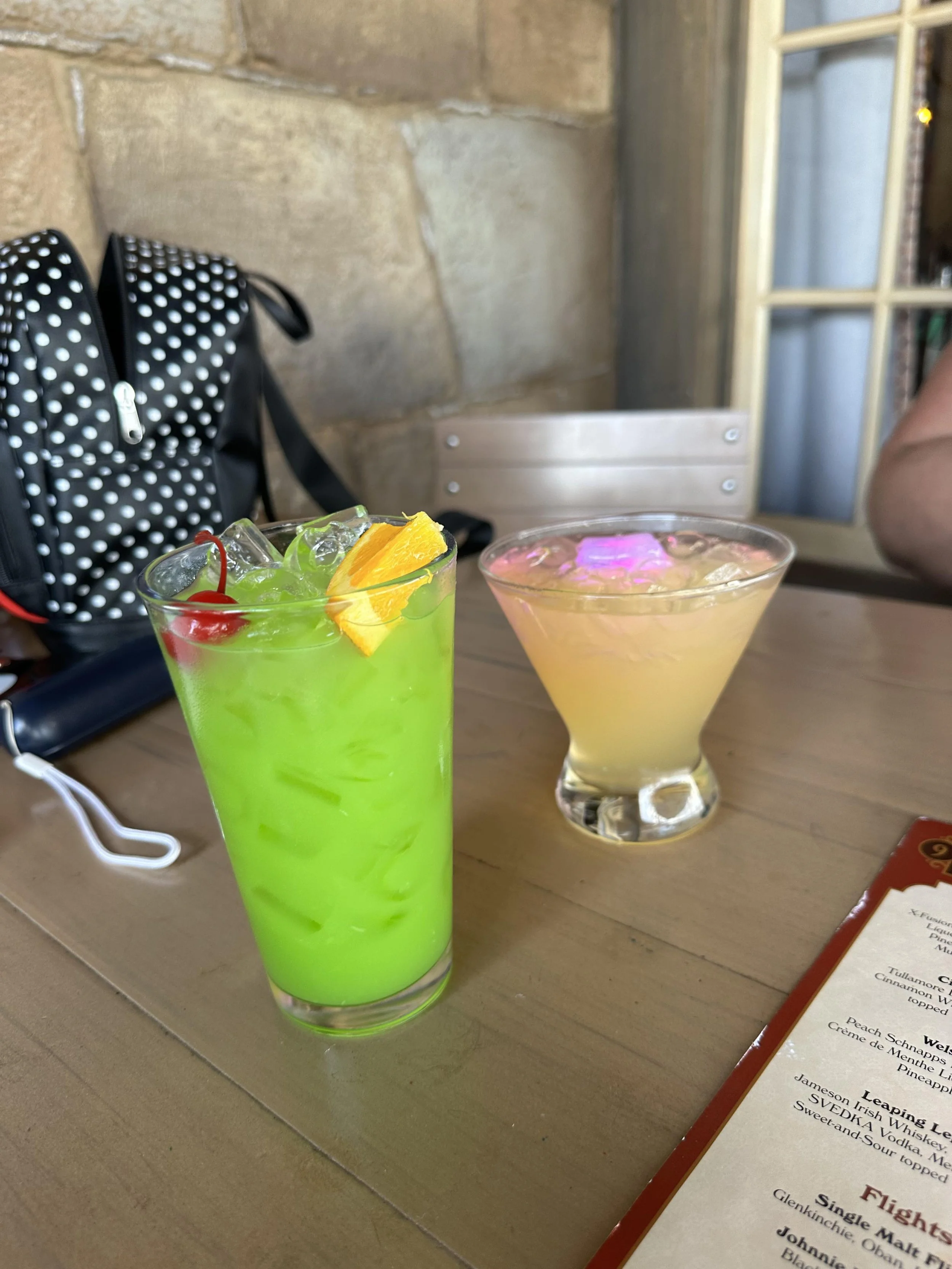 Two cocktails on a restaurant table, one bright green with ice and fruit garnishes, the other pinkish yellow in a cocktail glass.