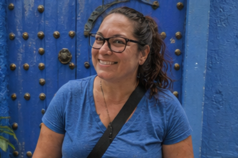 Smiling woman with glasses and curly hair, wearing a blue shirt, standing outdoors in front of a large blue door with decorative studs