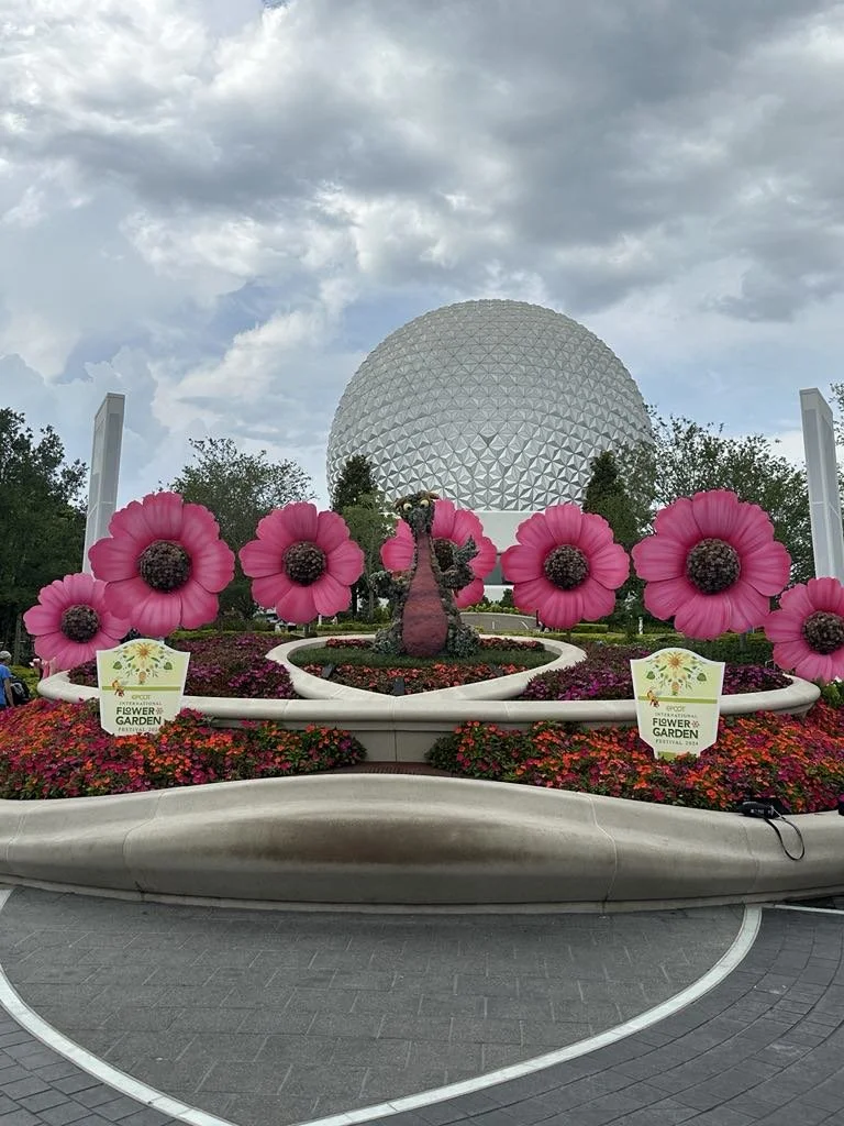Large flower bed with pink flowers and a pink dragon sculpture in the center, in front of a large geodesic sphere, with trees and cloudy sky in the background.