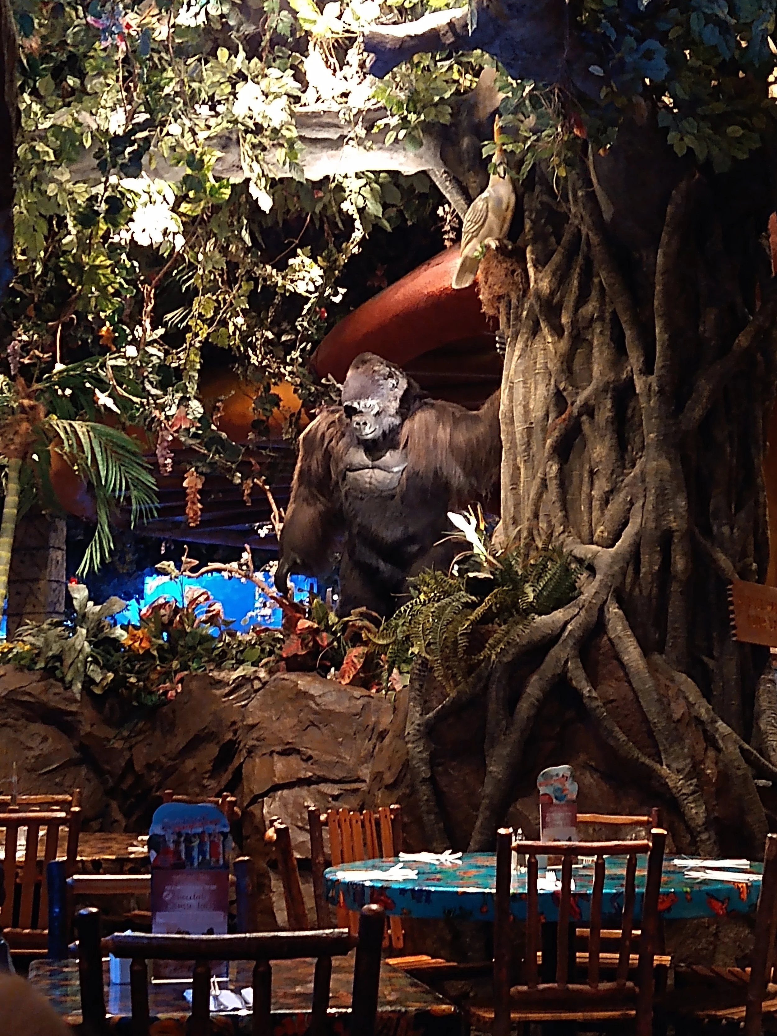 Indoor jungle-themed restaurant with a large gorilla statue, jungle plants, and animals like a bird on the tree trunk, surrounded by tables and chairs.