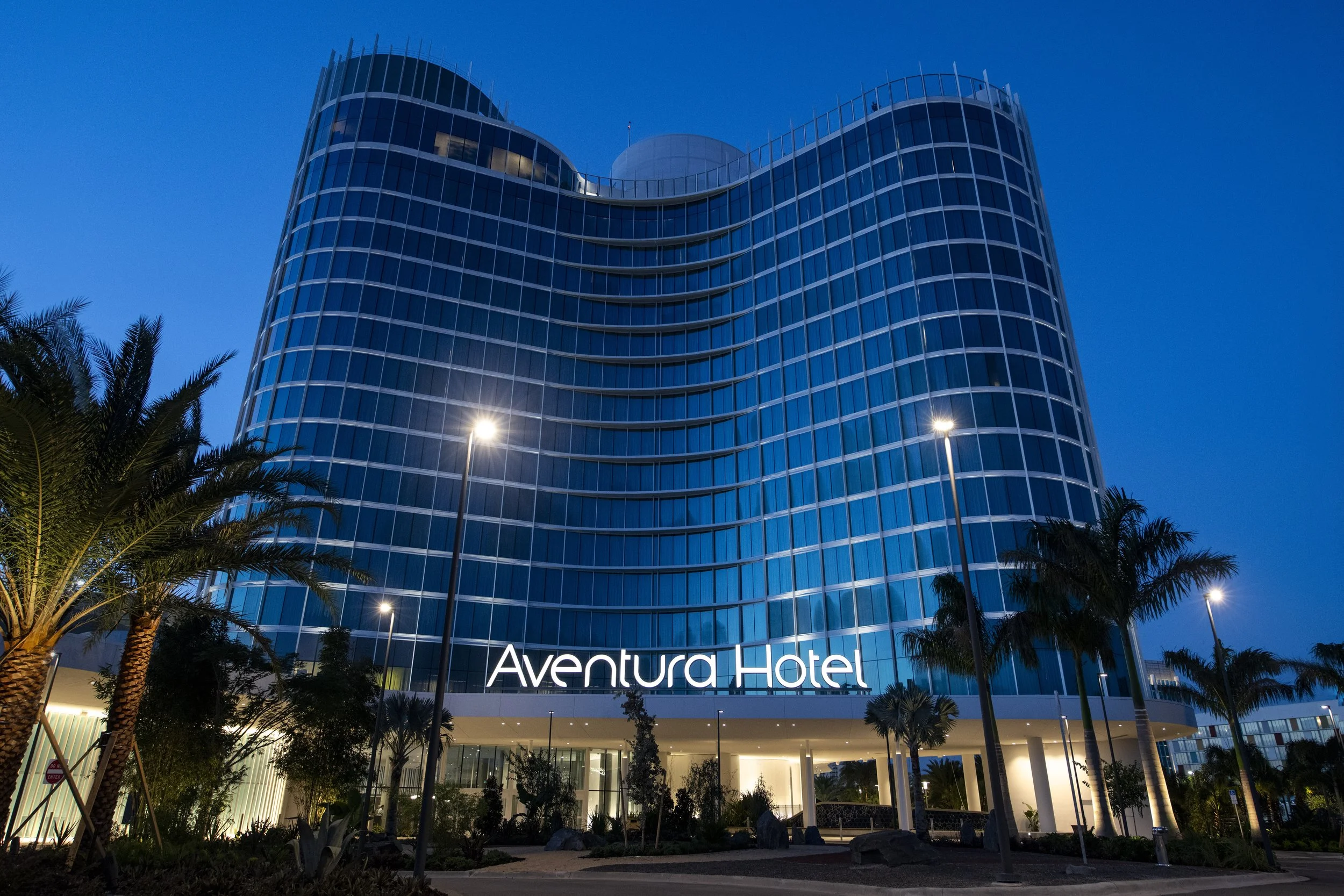 Night view of Aventura Hotel, a modern glass building with curved architecture, illuminated sign, surrounded by palm trees and streetlights.