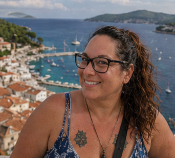 Woman with curly hair and glasses smiling, overlooking harbor with boats and coastline in the background.