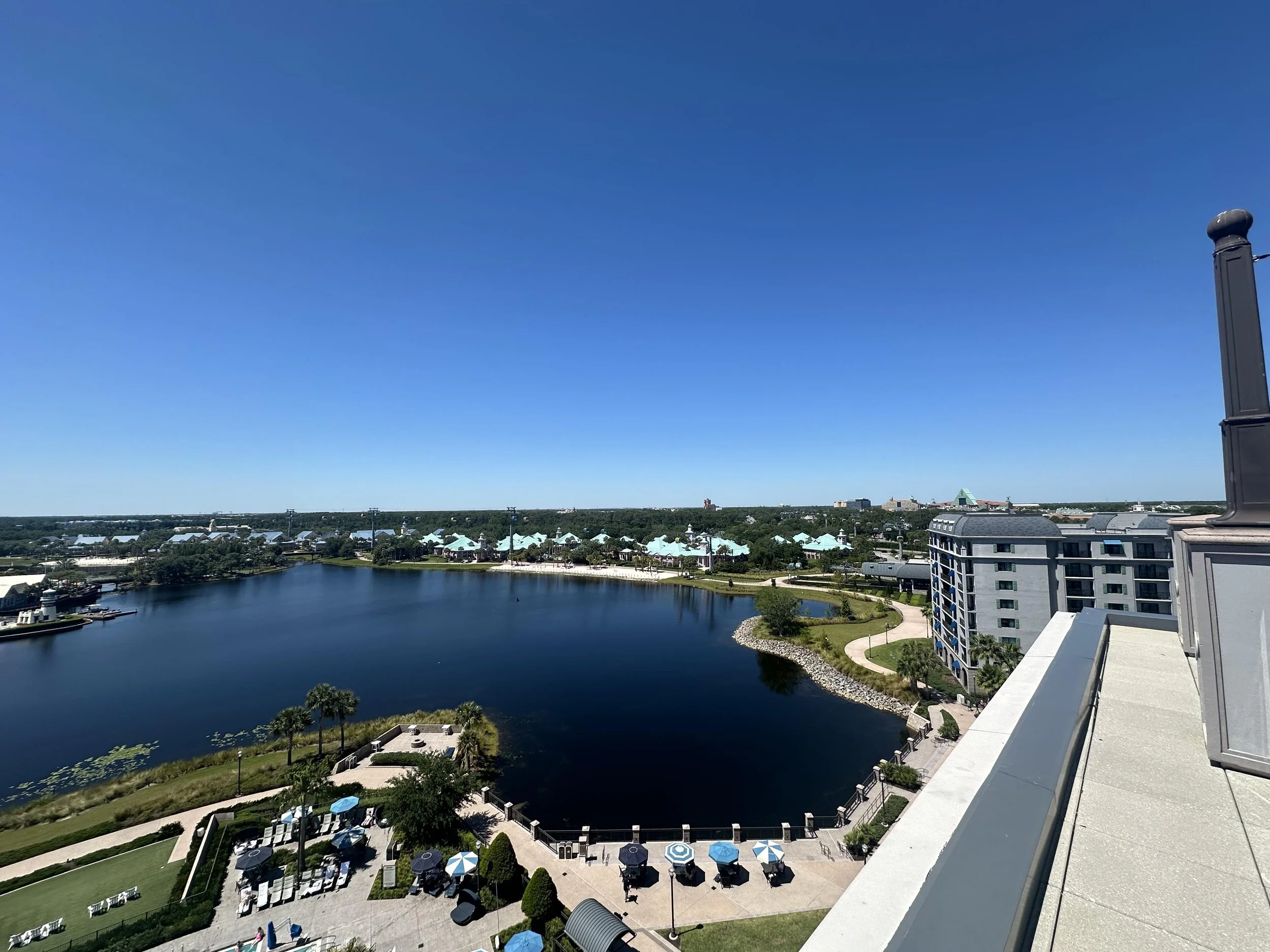 A high-angle view of a large body of water with a landscaped outdoor area, umbrellas, and lounge chairs, surrounded by tall modern buildings and a clear blue sky.