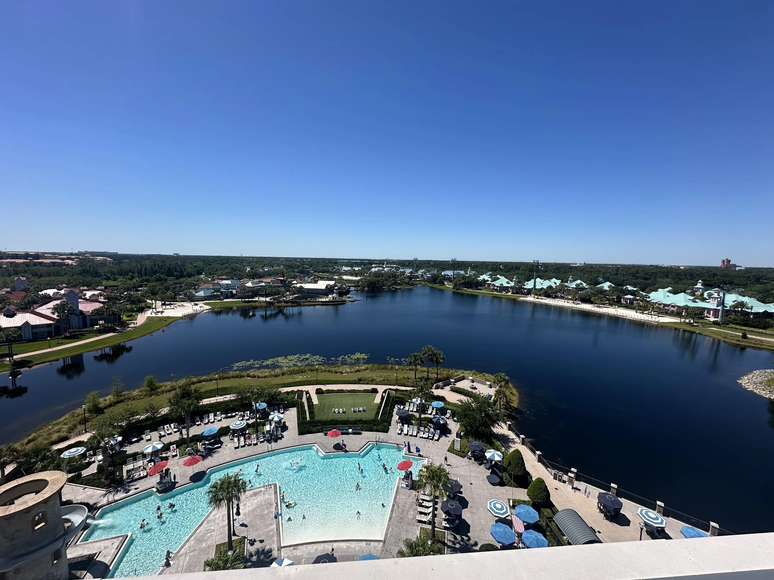 Aerial view of a swimming pool complex with umbrellas and lounge chairs, overlooking a calm lake with surrounding greenery and buildings under a clear blue sky.