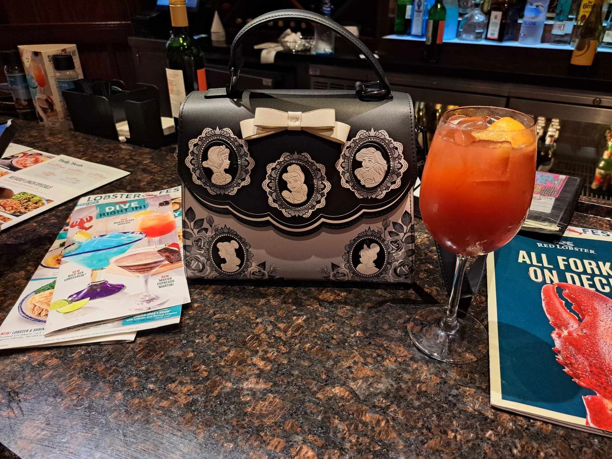 A cocktail drink with ice and an orange slice in a wine glass, a black and white purse with portrait illustrations and a bow on top, and several menus and pamphlets on a bar counter.