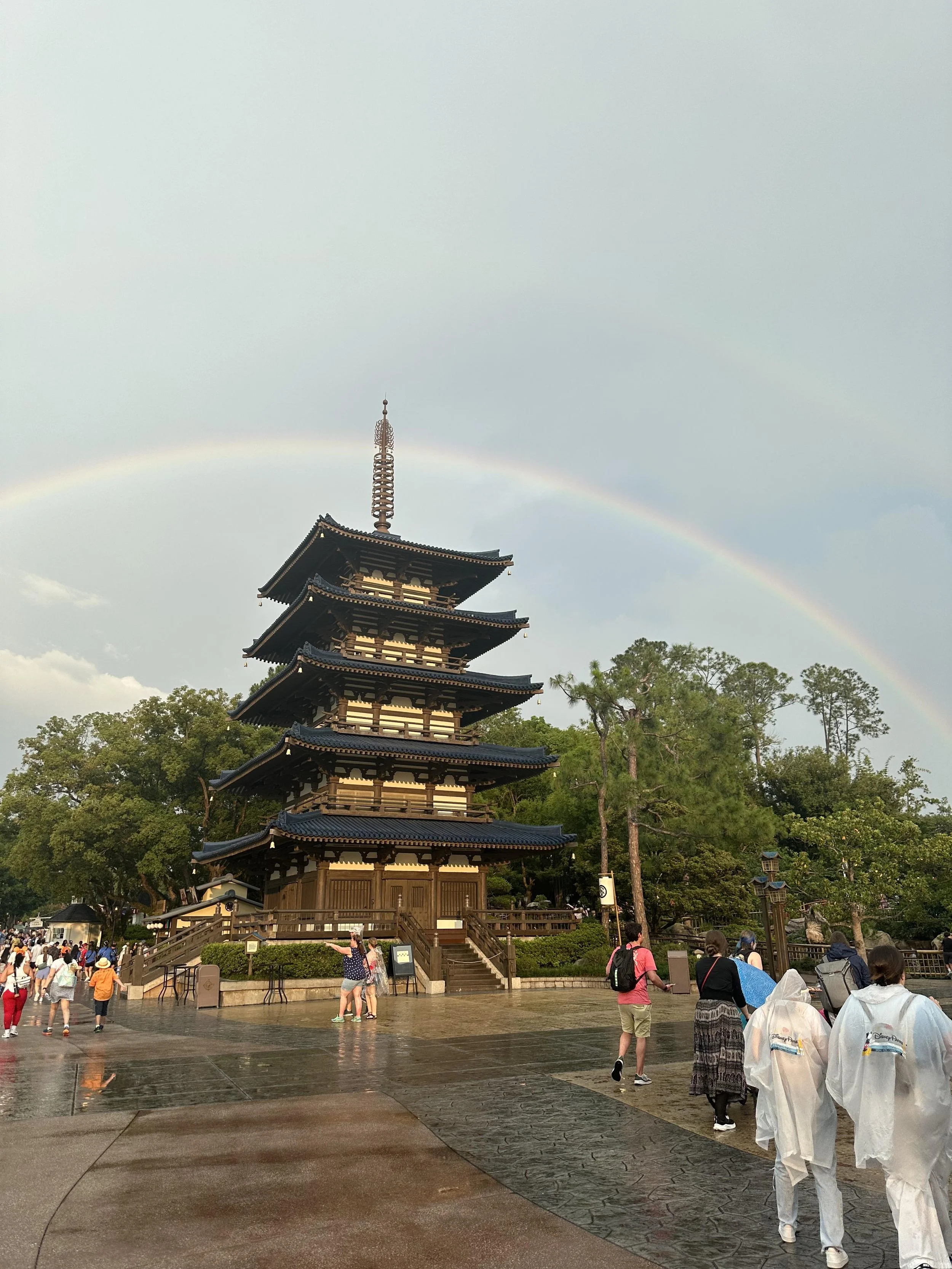 A traditional Japanese five-story pagoda with a rainbow in the sky above, surrounded by trees and visitors at a park or temple.
