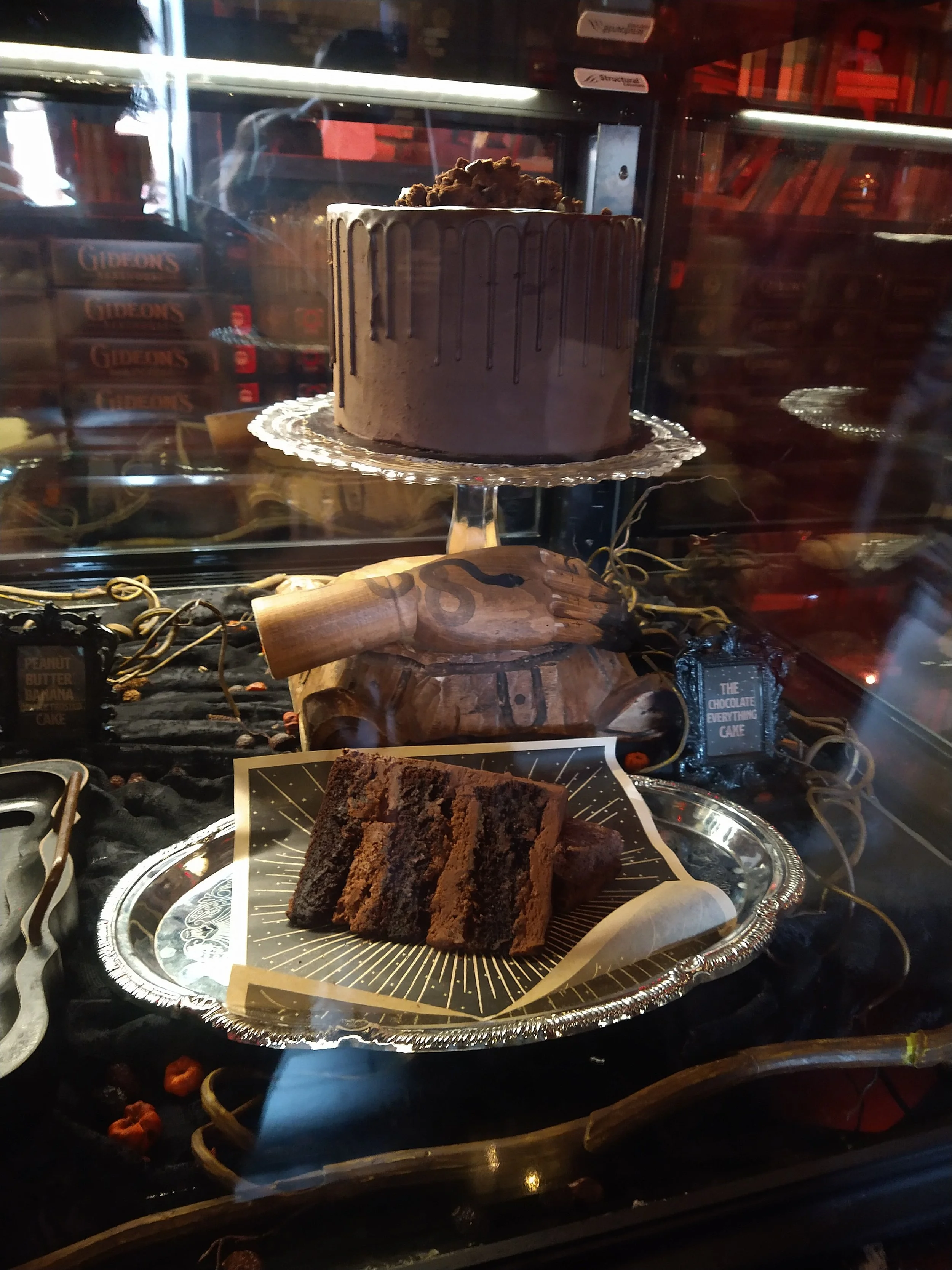 Chocolate cake with a drizzle of chocolate ganache on top, displayed behind glass in a bakery case, with slices of chocolate cake on a silver tray in front.