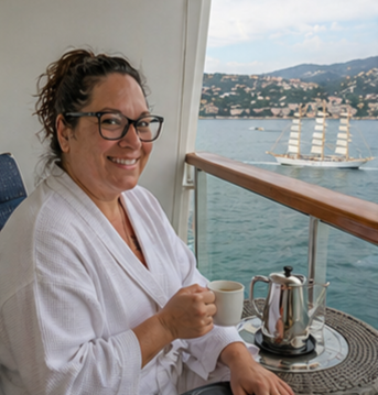 Woman in glasses smiling and holding a cup of coffee or tea on a boat, with a scenic waterfront and sailing ship in the background.