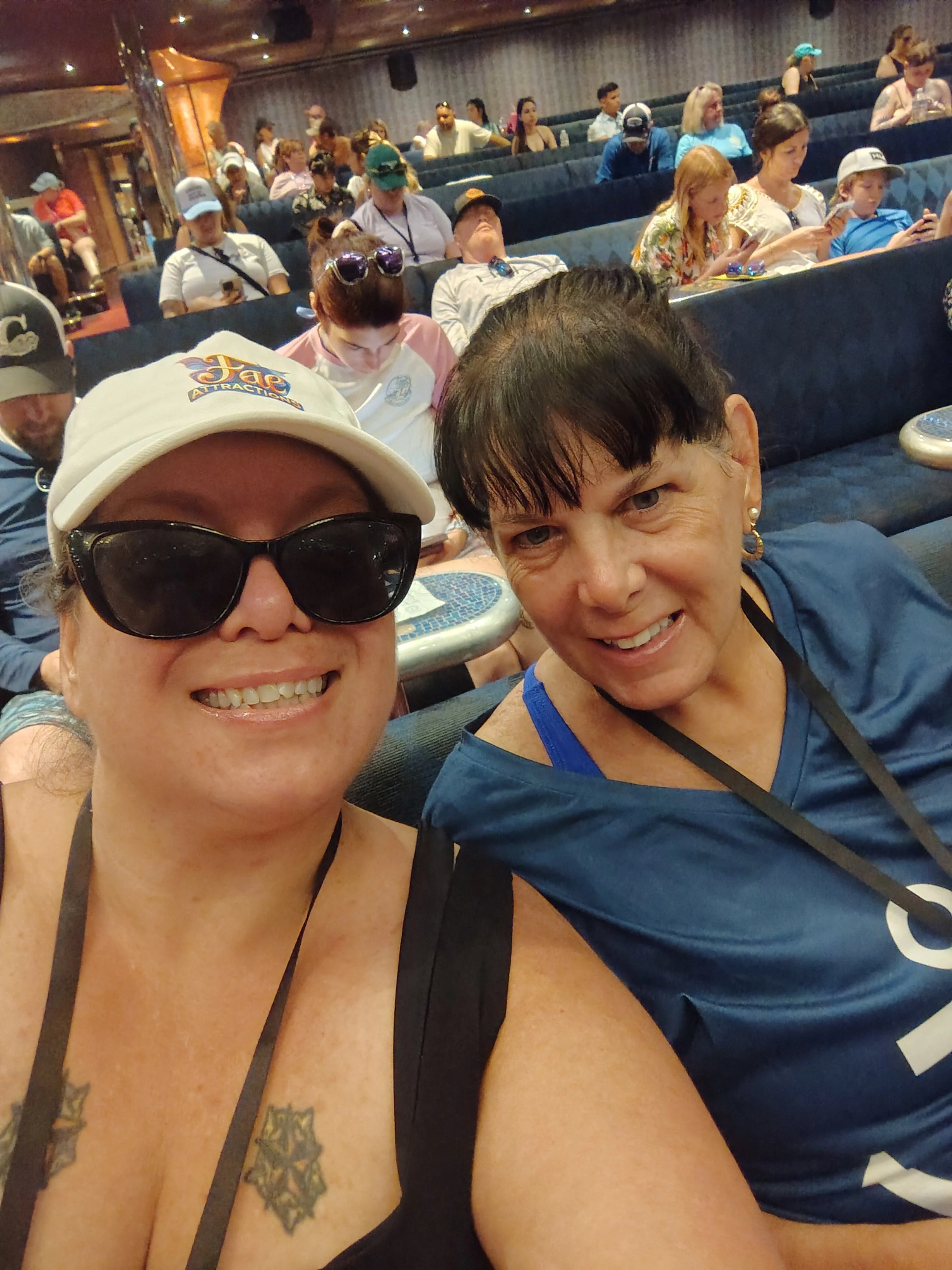 Two women smiling, taking a selfie at a crowded indoor event with many people seated behind them.