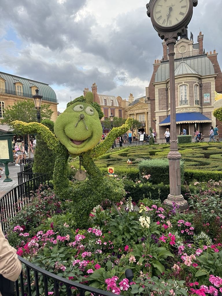 Topiary sculpture of a green character with glasses and a big smile, surrounded by pink and purple flowers in a garden, with a large clock and a European-style building in the background.