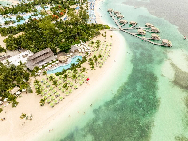 Aerial view of a tropical beach resort with a sandy shoreline, numerous beach chairs and umbrellas, a swimming pool surrounded by palm trees, lush greenery, overwater bungalows connected by a wooden pier extending into the turquoise ocean.
