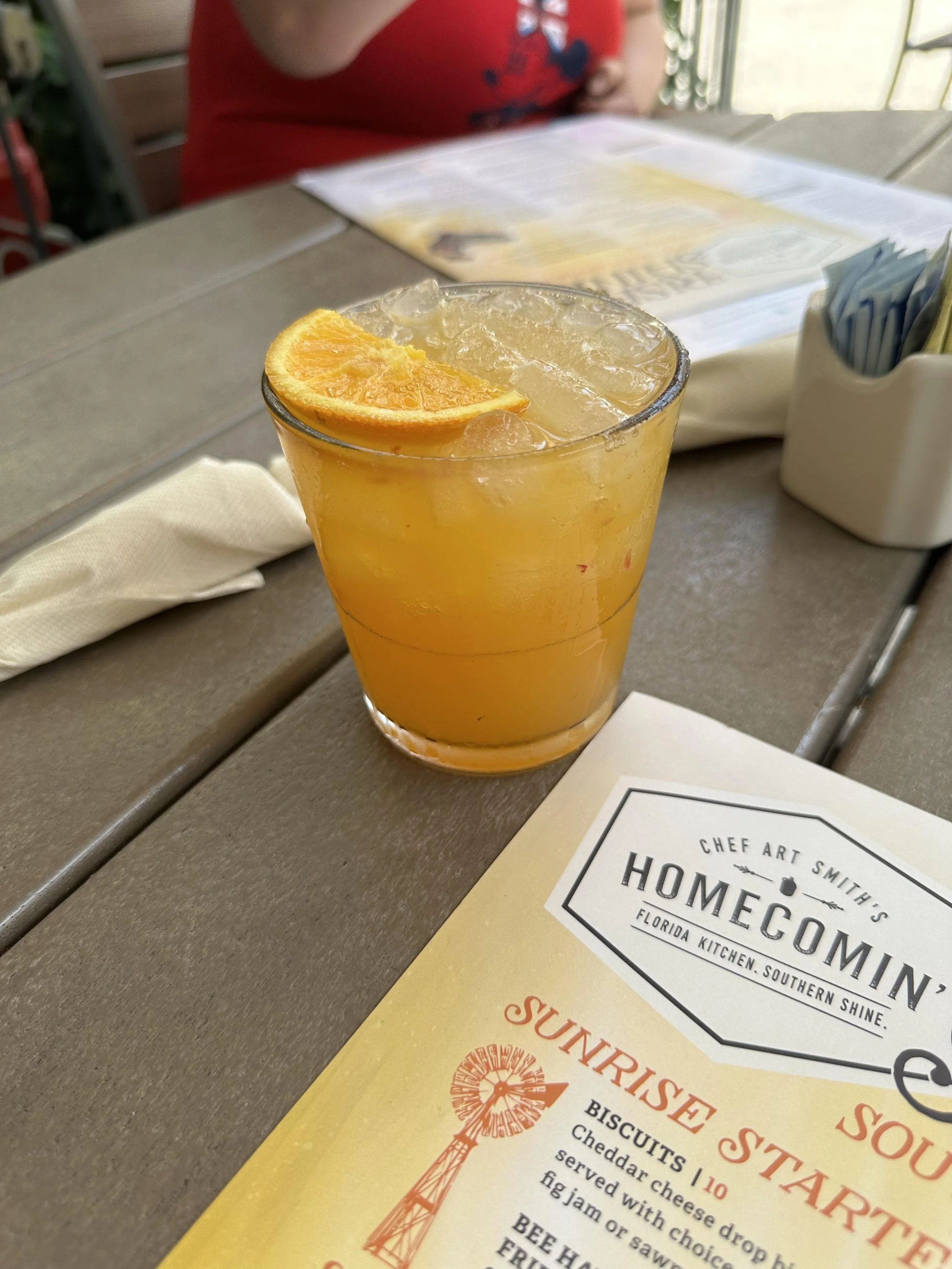 A glass of orange juice with ice cubes and an orange wedge on top, placed on a table at a restaurant. A menu and some utensils are also visible on the table.