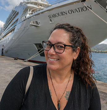 A woman with curly hair, glasses, and a black shirt smiling at the camera near a dock, with a large white cruise ship named 'Seabourn Ovation' in the background.