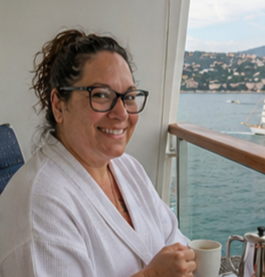Smiling woman with glasses holding a cup on a boat deck, with water and mountains in the background.