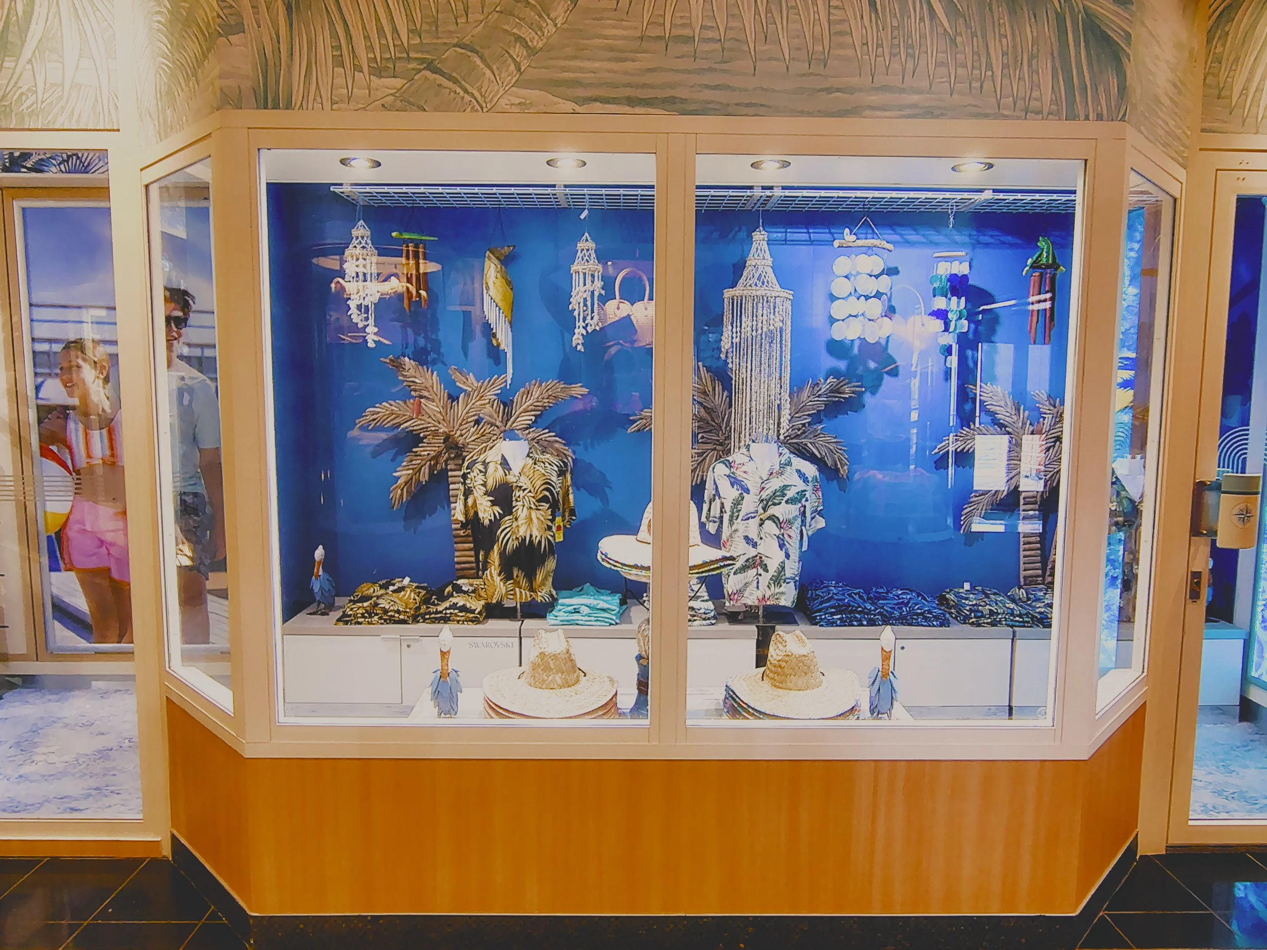 Display case with tropical-themed clothing, hats, and decor, featuring mannequins with palm leaf accessories and hanging shell and beaded decorations, set against a blue background.