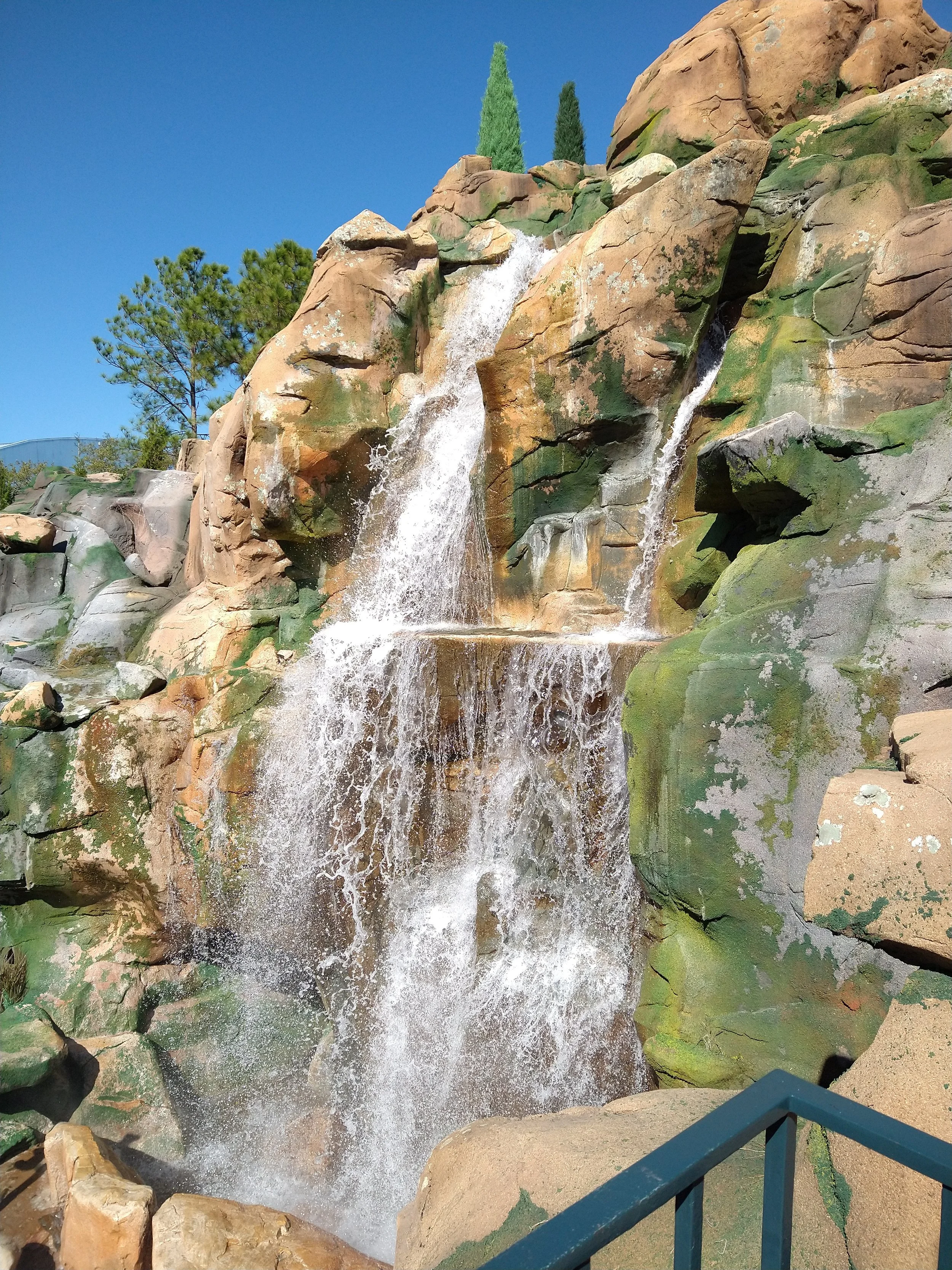 Waterfall cascading over rocky terrain with green moss and trees in a sunny outdoor setting.