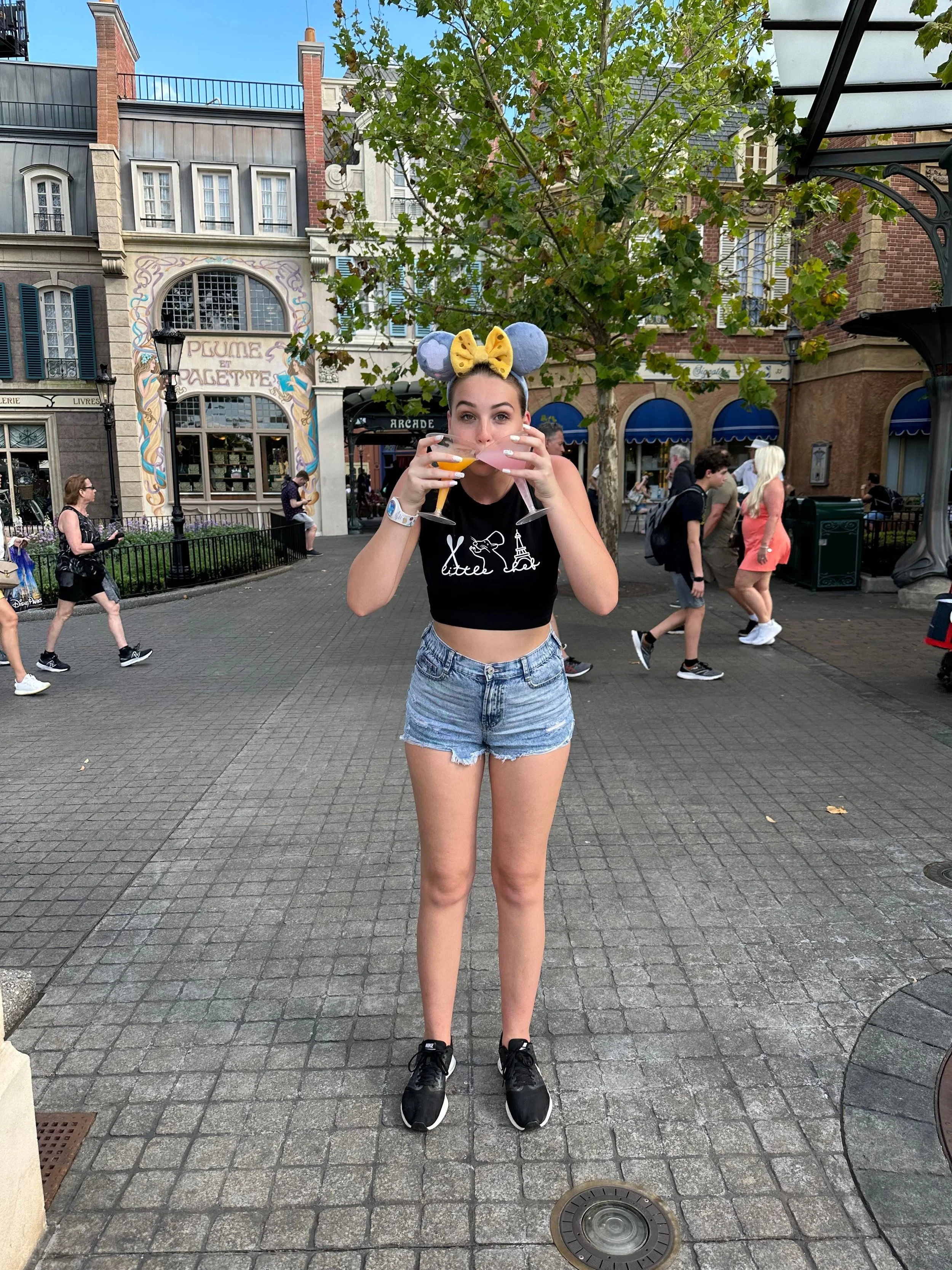 Young woman standing on a cobblestone street, holding a drink in each hand, wearing Mickey Mouse ears headband with yellow bow, black crop top, and denim shorts. In the background, there are other people, storefronts, and a decorative building, sugge