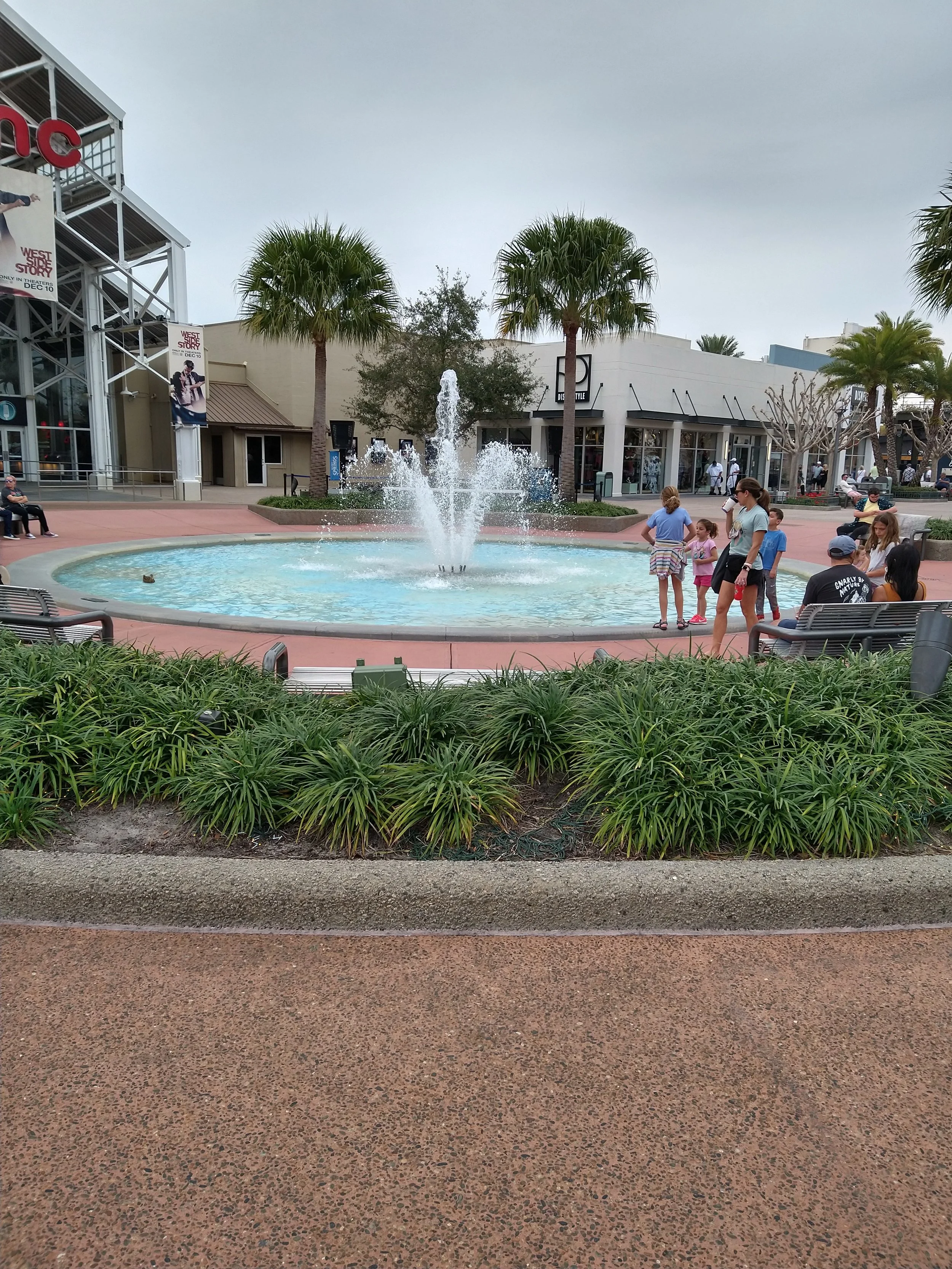 Public square with a fountain, people sitting on benches, and children standing near the fountain. Storefronts and palm trees in the background under a cloudy sky.