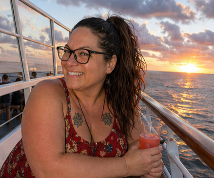 Woman with glasses smiling on a boat during sunset, holding a drink.