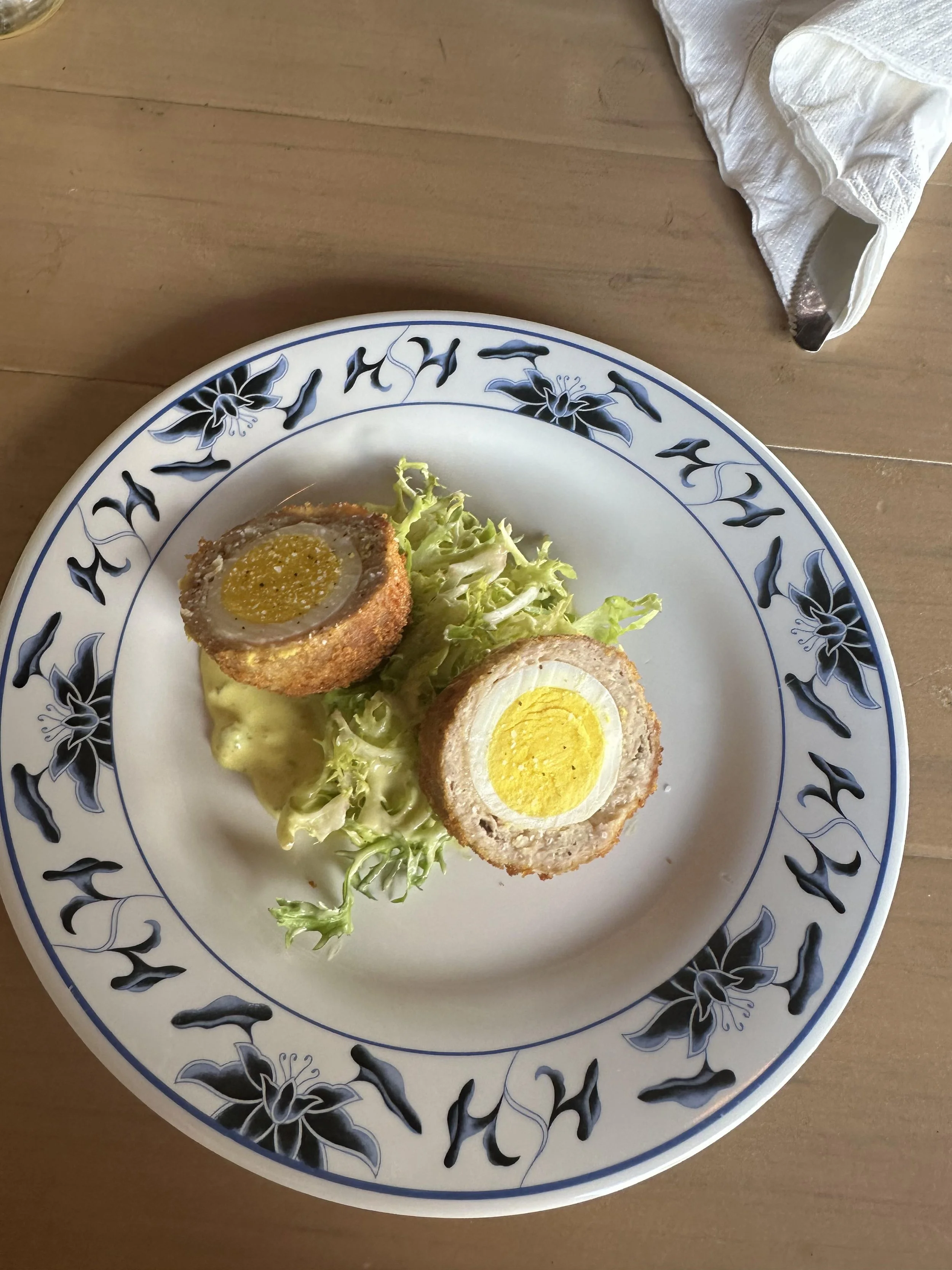 Plate with a Scotch egg cut in half, served with a leaf of lettuce and mustard sauce on a decorative blue and white plate.