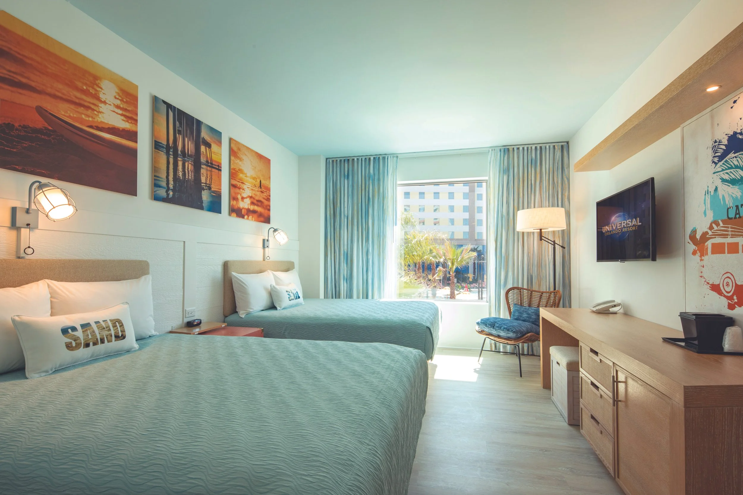 A hotel room with two beds, wall art of a sunset and pier, a window showing a building exterior, a TV on the wall, a chair, and a wooden desk.