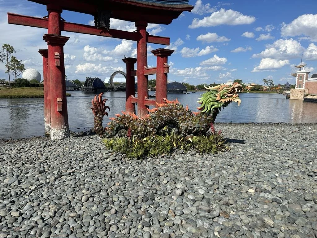 A decorative dragon sculpture next to a red torii gate outdoors with water, trees, and amusement park structures in the background.